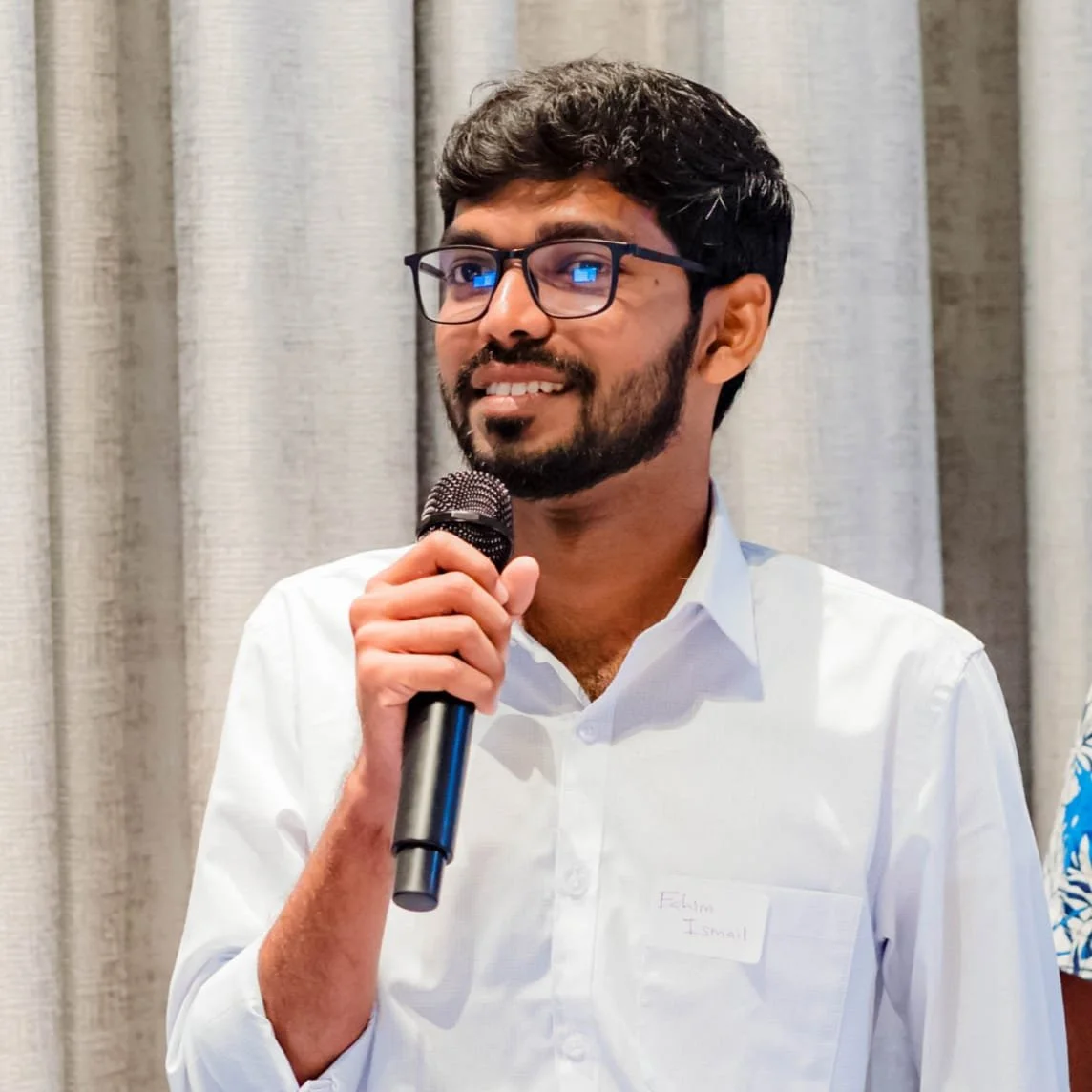 Fahim - outreach and translation consultant. A man with glasses, a beard, and a mustache holding a microphone, smiling, in front of a gray curtain.