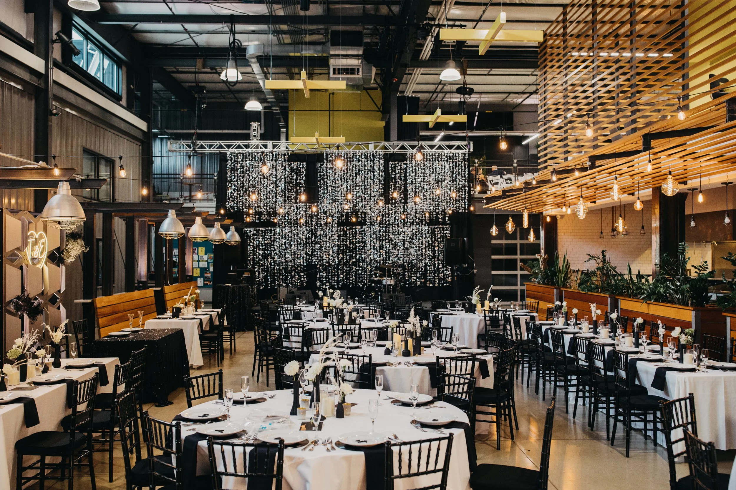 Interior of a decorated banquet hall with black chairs, round tables with white tablecloths, and hanging string lights and chandeliers.