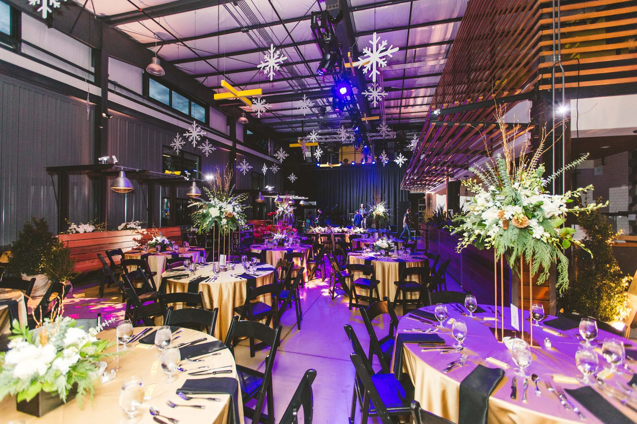 Indoor event space decorated with floral centerpieces, hanging snowflake decorations, and purple lighting, with tables set for a formal occasion.