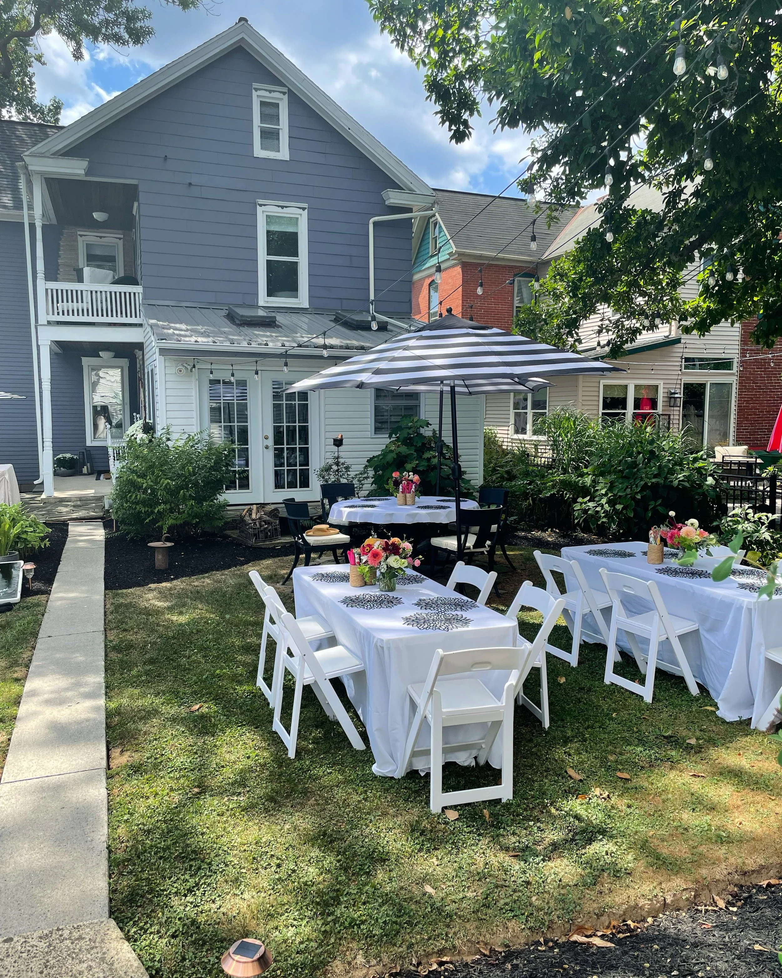 Outdoor garden party setup with tables covered in white tablecloths, floral centerpieces, and an umbrella, in a backyard with houses and greenery.