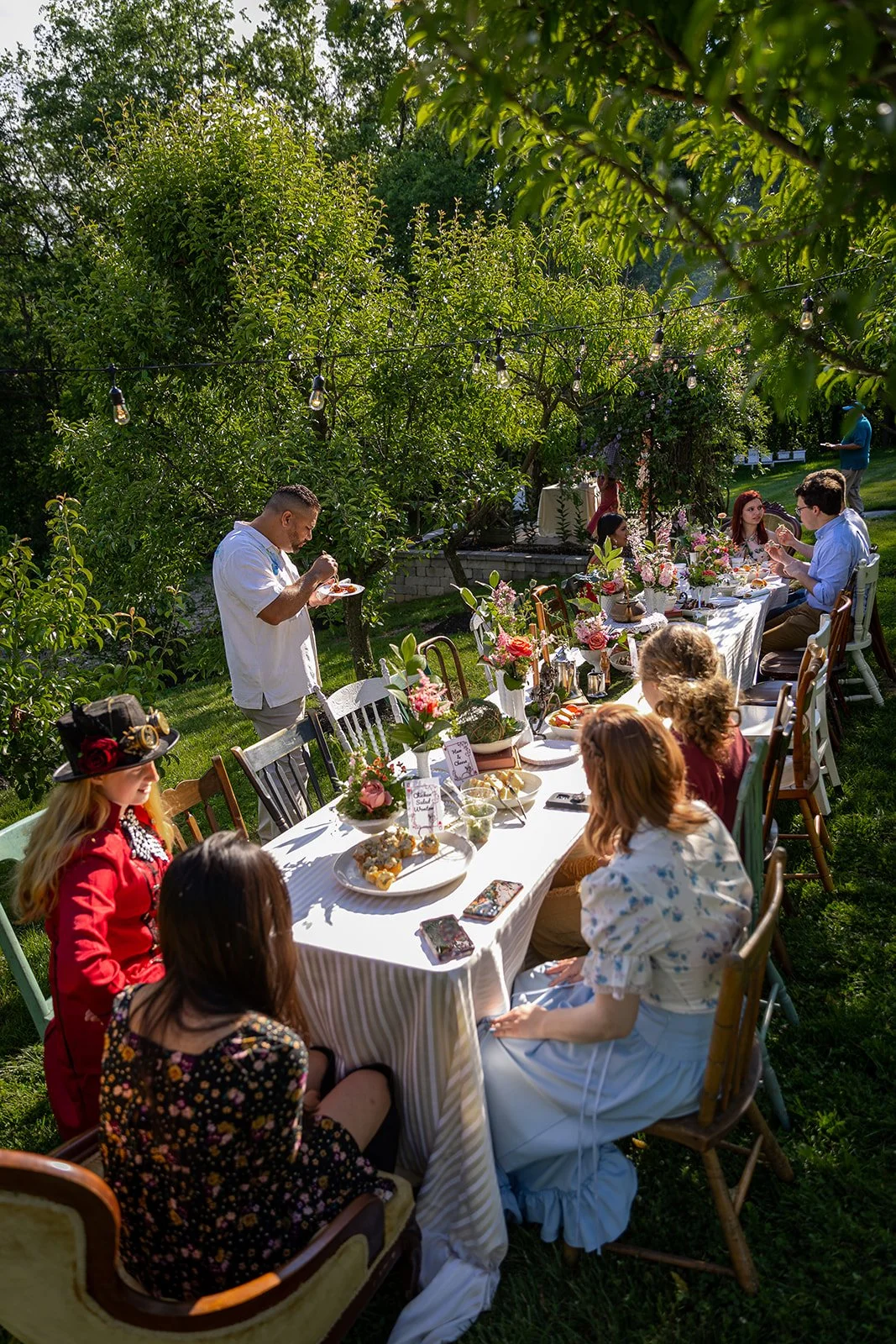 People gather around a long outdoor dining table decorated with flowers and greenery, enjoying a meal in a lush garden during daylight, with string lights hanging overhead.