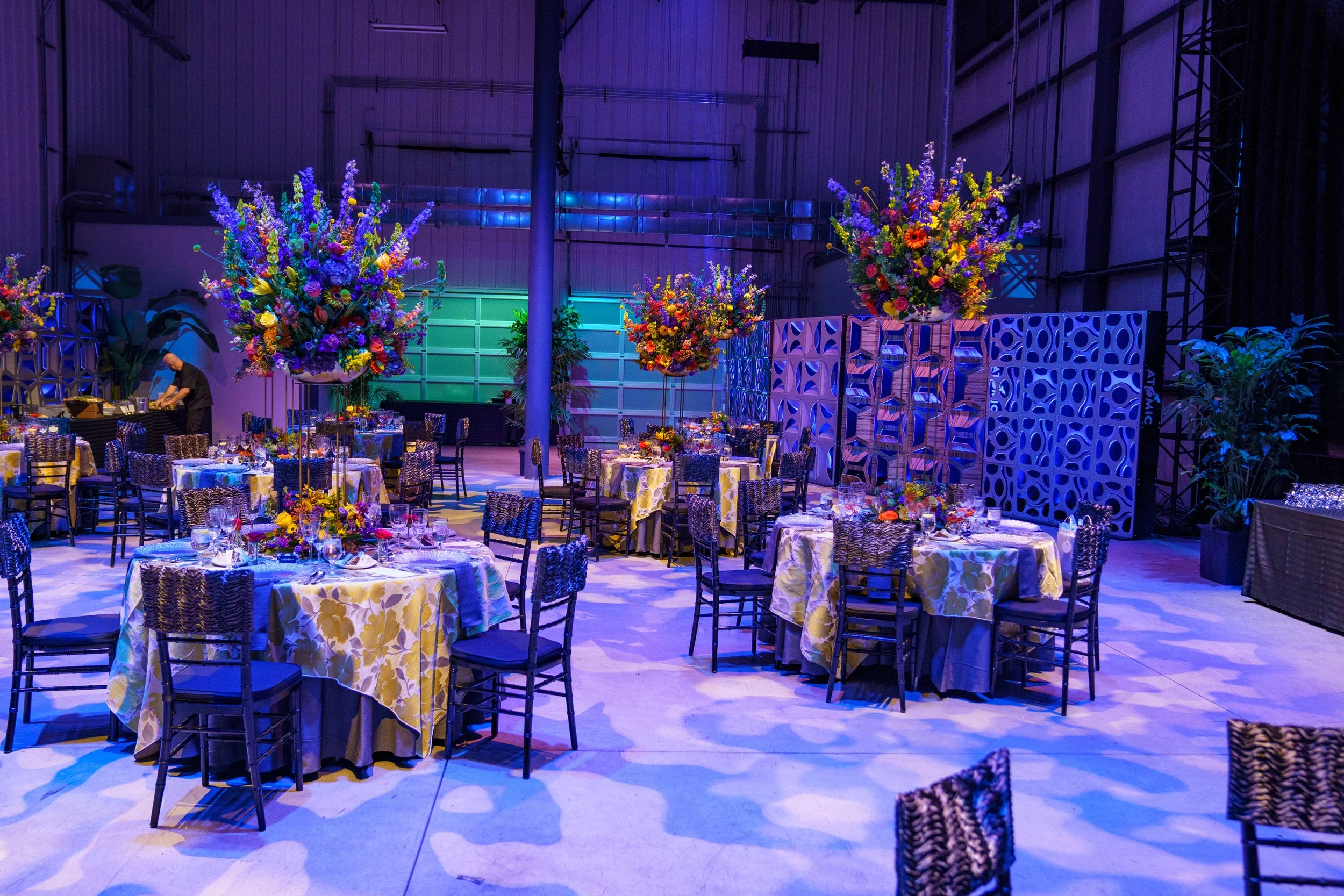 Event hall decorated with floral centerpieces on round tables covered with yellow floral tablecloths, set for a formal event, with blue lighting and decorative panels in the background.