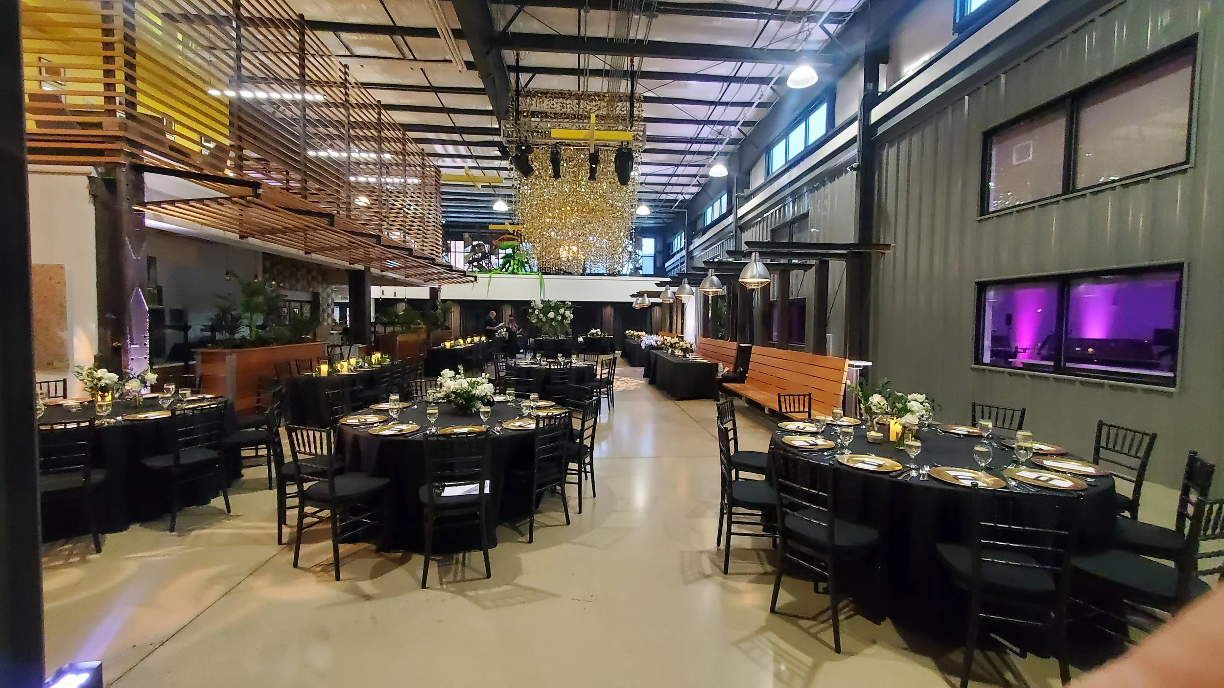 Elegant event space with round tables covered in black tablecloths, decorated with white floral centerpieces and gold-rimmed plates, set for a formal occasion. The room features a large, ornate chandelier, modern pendant lighting, a wooden balcony st