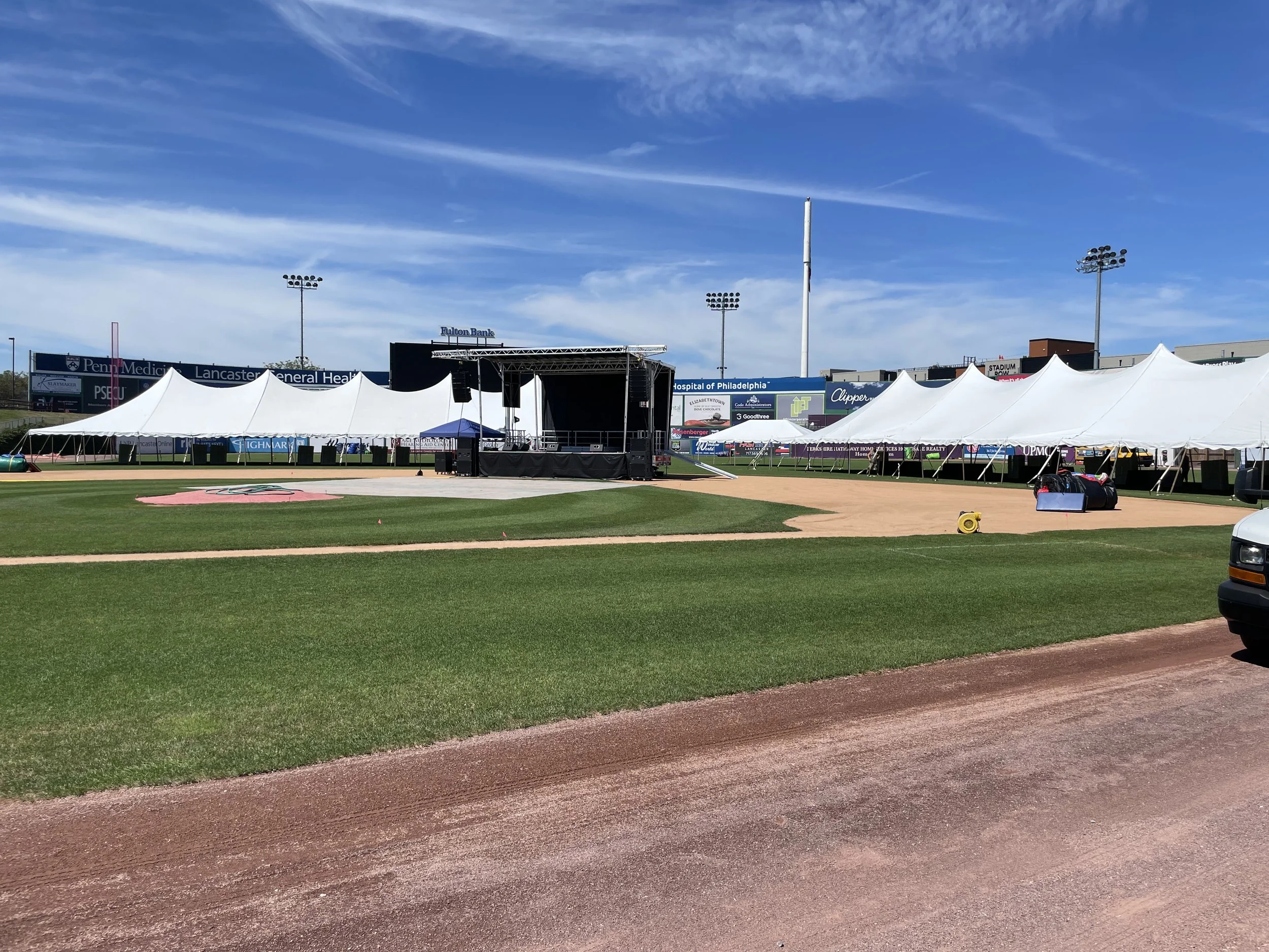 A baseball stadium field under a blue sky with scattered clouds. There are white tents set up along the outfield, a stage with speakers near the center, and multiple tall light poles. The field has green grass and a dirt track surrounding the infield