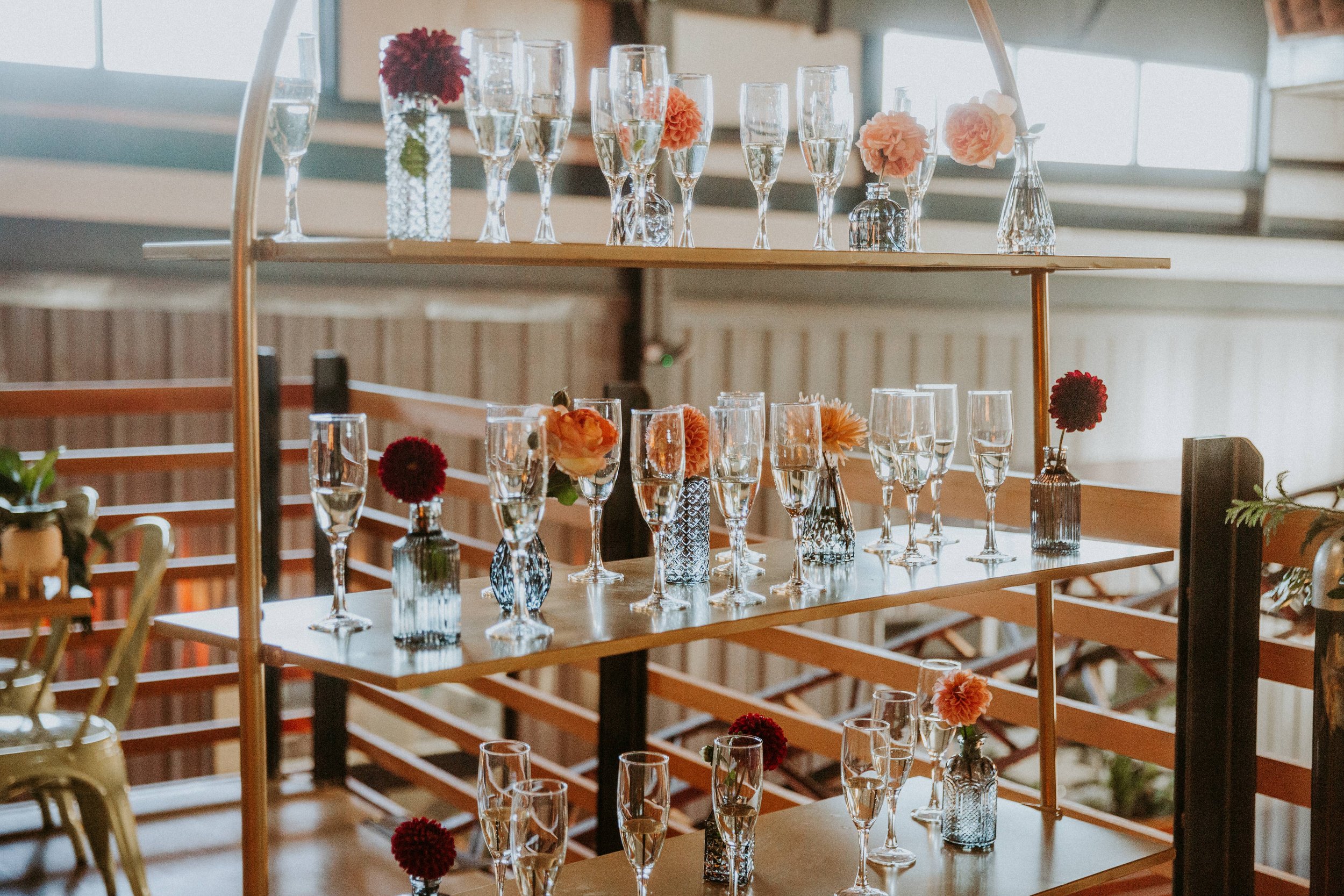 Display of champagne glasses and small flower vases with pink and red flowers on a multi-tiered wooden and metal table in a well-lit indoor setting.