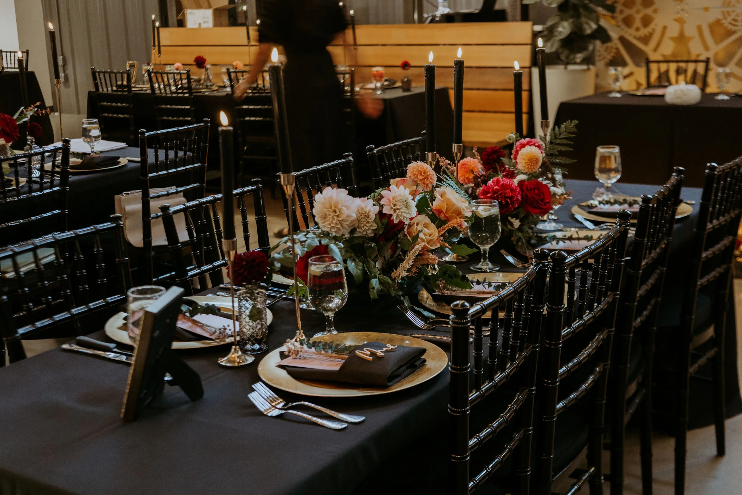 Elegant banquet table decorated with a floral centerpiece of pink, white, and red flowers, black candles in gold holders, set with black napkins, gold chargers, and glassware, in a dimly lit room.