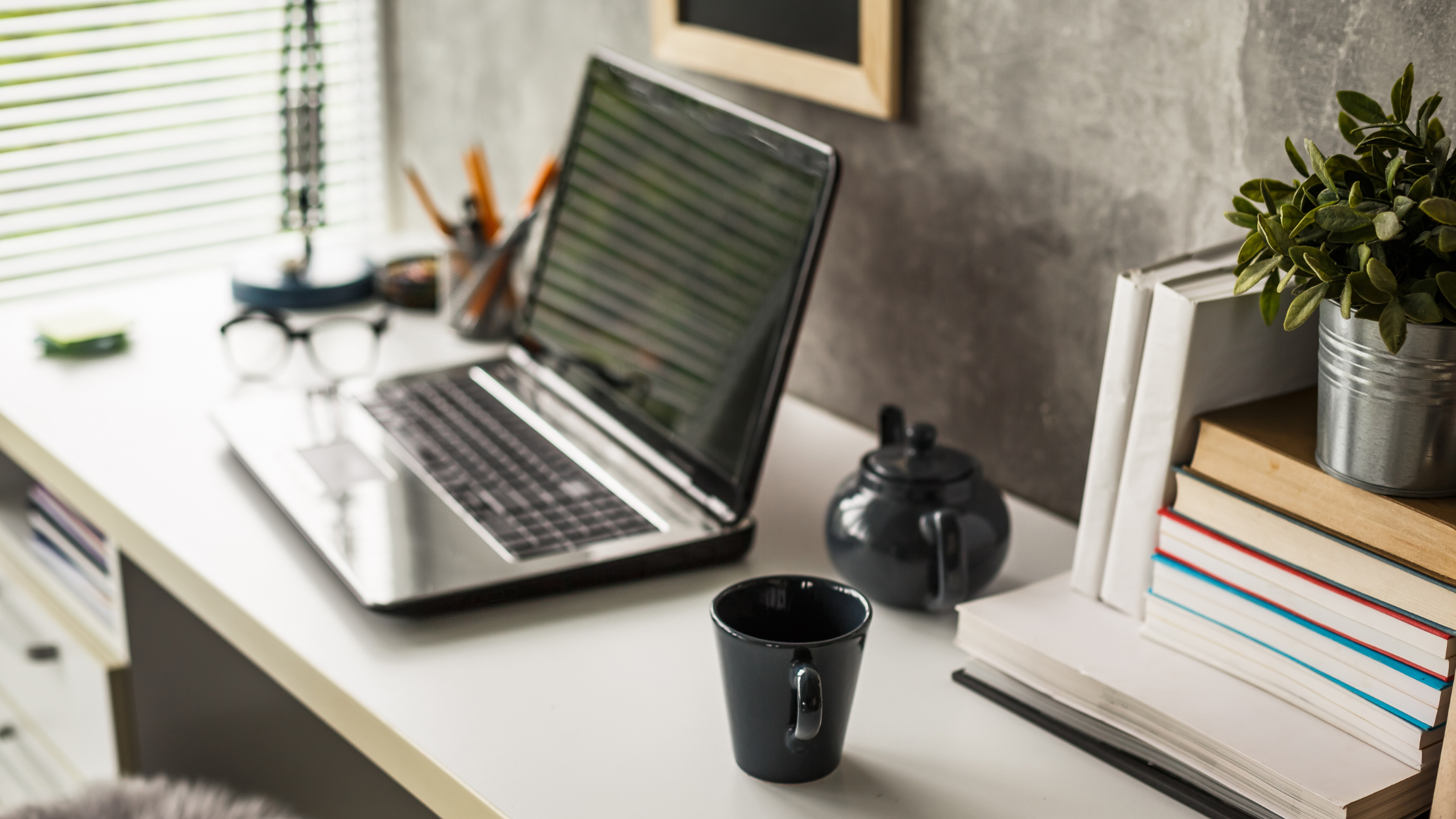 A modern workspace with a white desk, an open laptop, black coffee mug, black teapot, books, a potted plant, and glasses near a window with blinds.