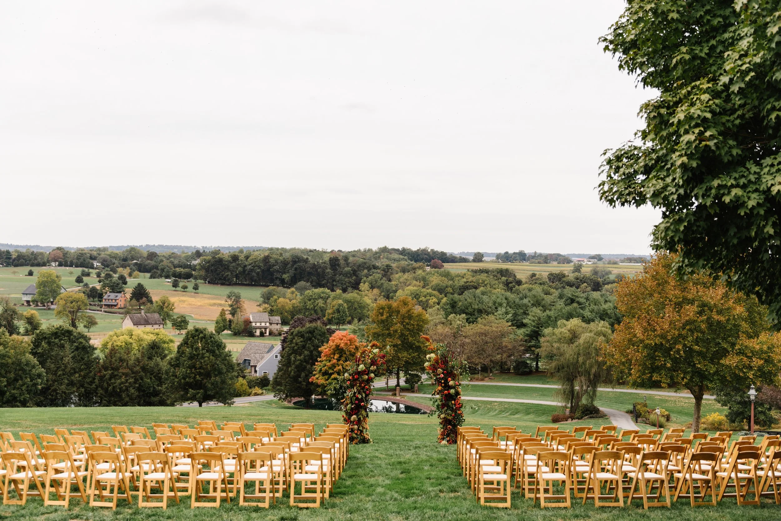 Outdoor wedding ceremony setup with rows of yellow chairs facing a floral arch in a scenic green landscape with trees and hills.