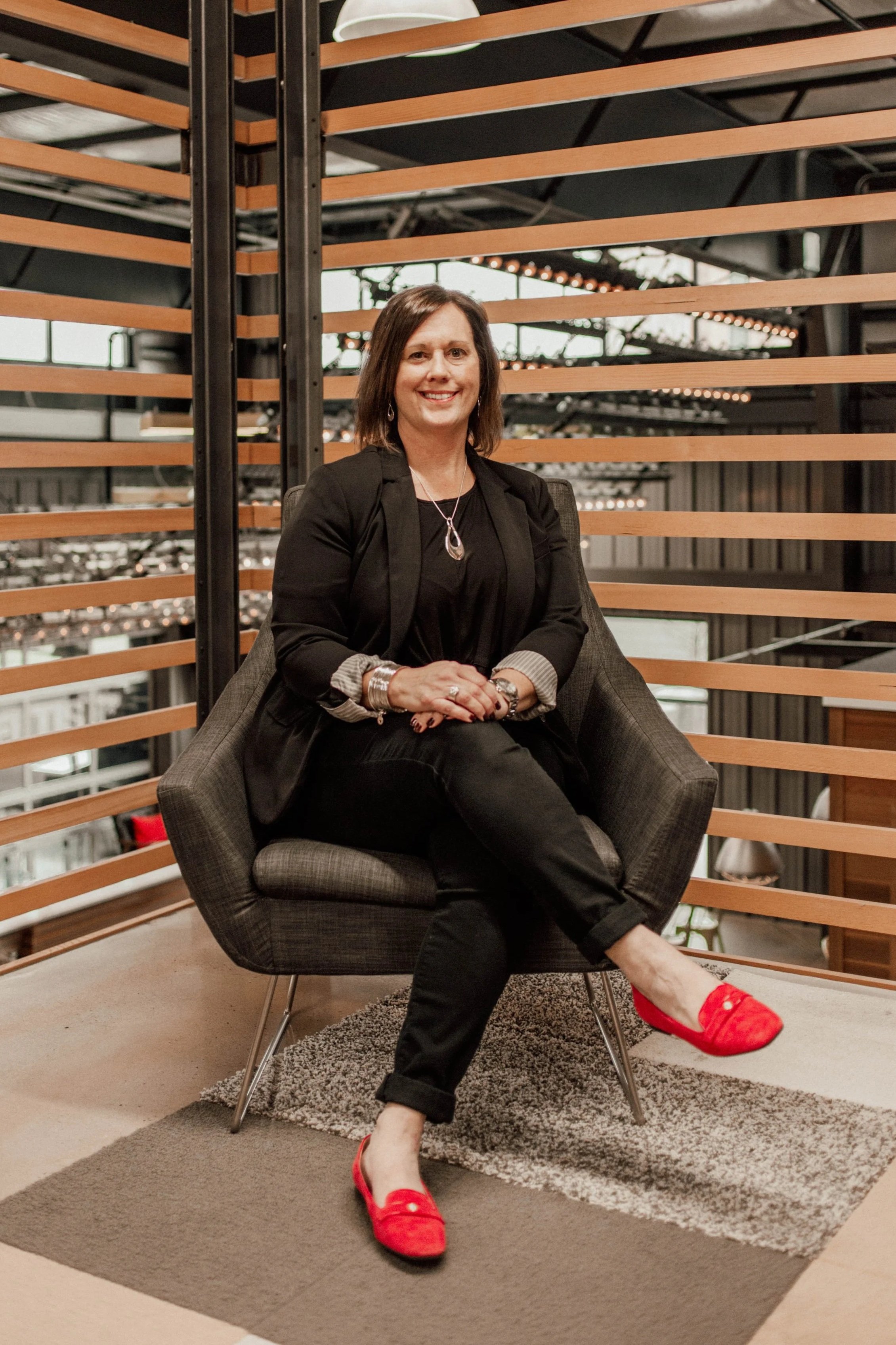 A woman sitting in a modern lounge area, wearing a black blazer, black pants, bright red flats, and accessories, with a wooden and metal decor background.