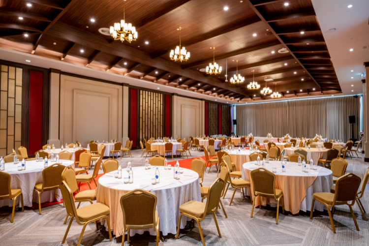 Elegant banquet setup with round tables covered in white tablecloths, surrounded by beige chairs, in a large decorated event hall with wood-paneled ceiling and chandeliers.