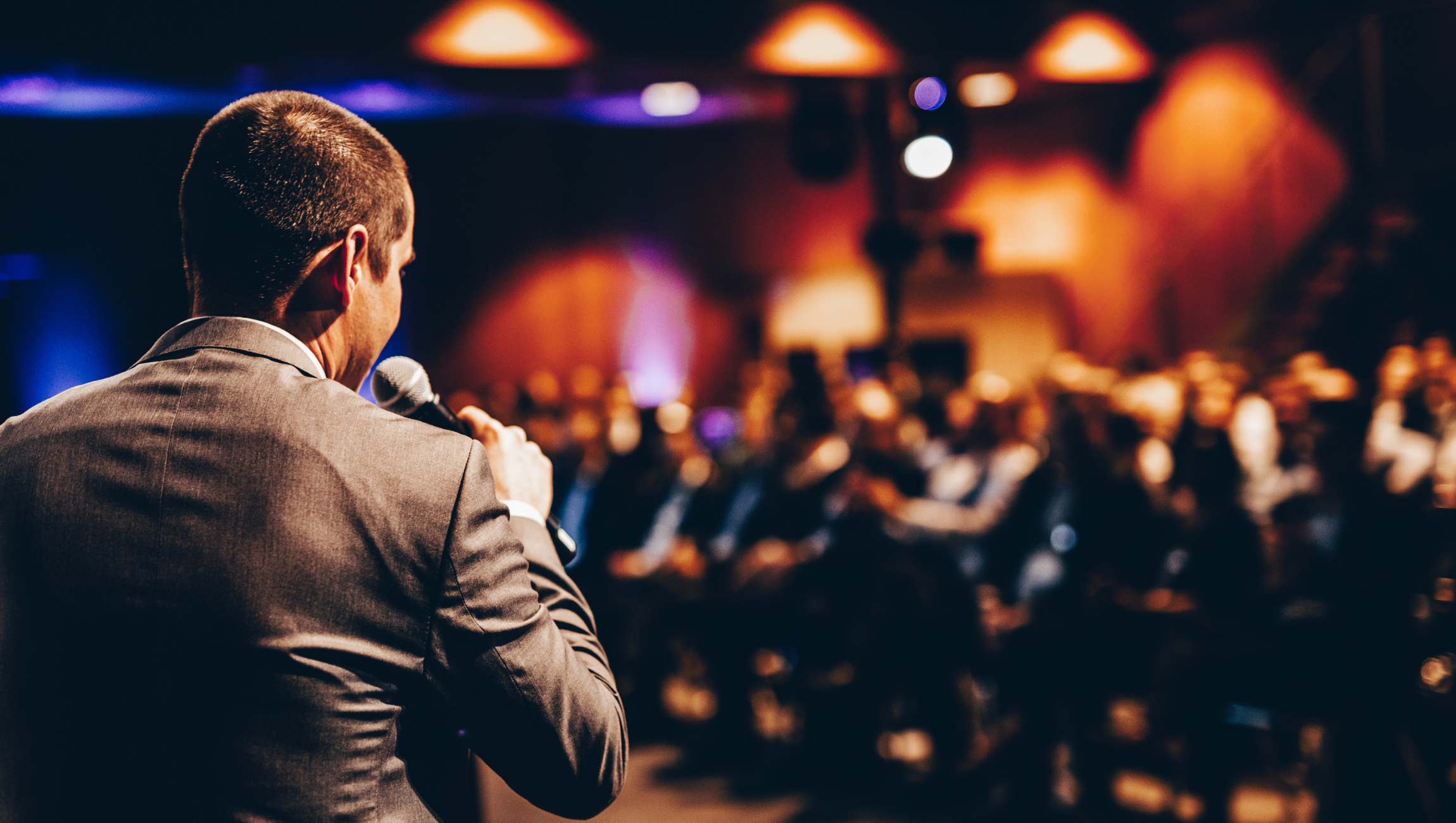 Man in a gray suit holding a microphone, speaking at an event in a large, dimly lit room filled with seated attendees.