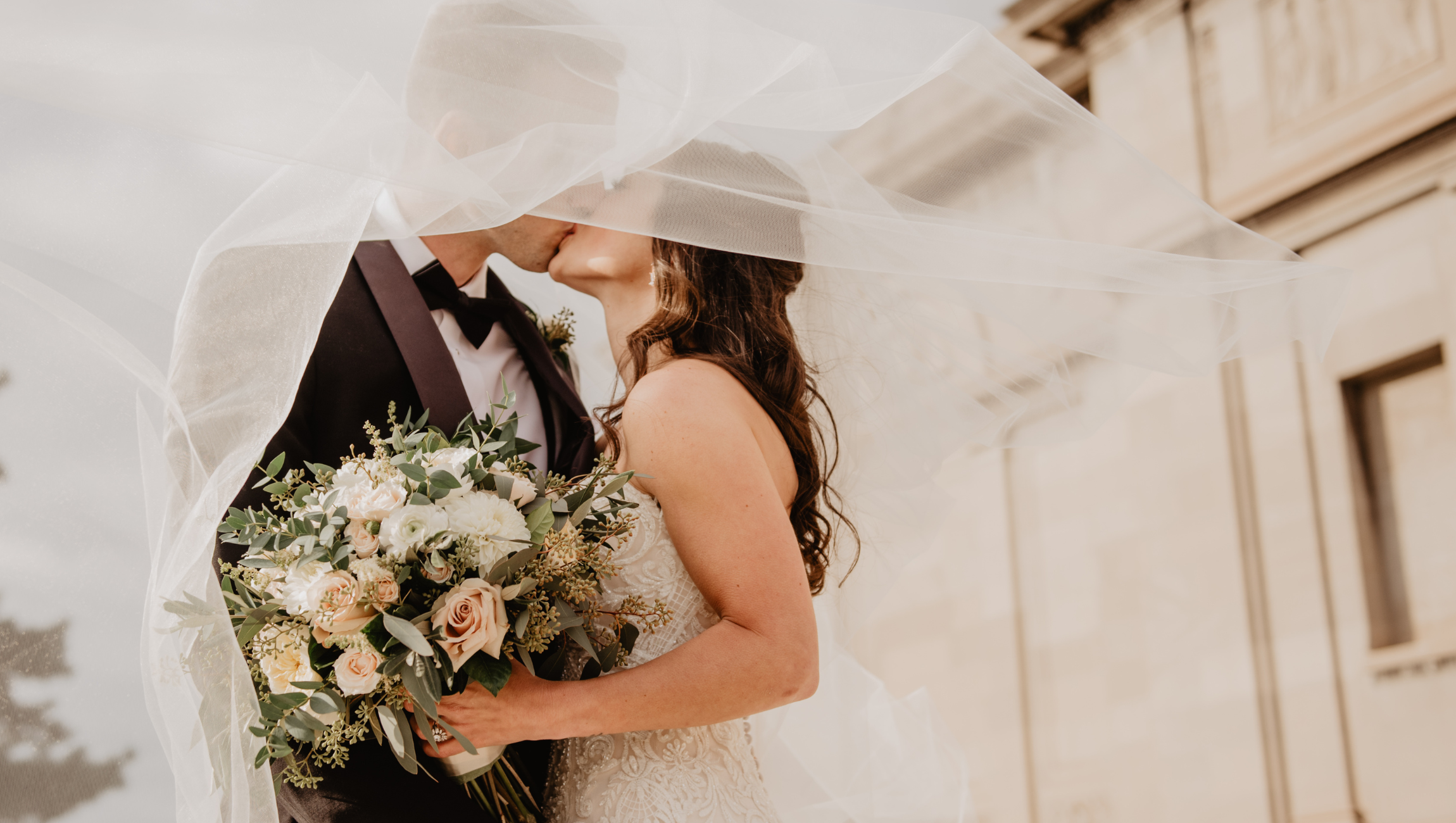 A bride and groom are about to kiss, partially obscured by the bride's veil. The bride holds a bouquet of white and blush roses, greenery, and other flowers. The groom wears a black tuxedo with a bow tie, and the background features a light-colored building.