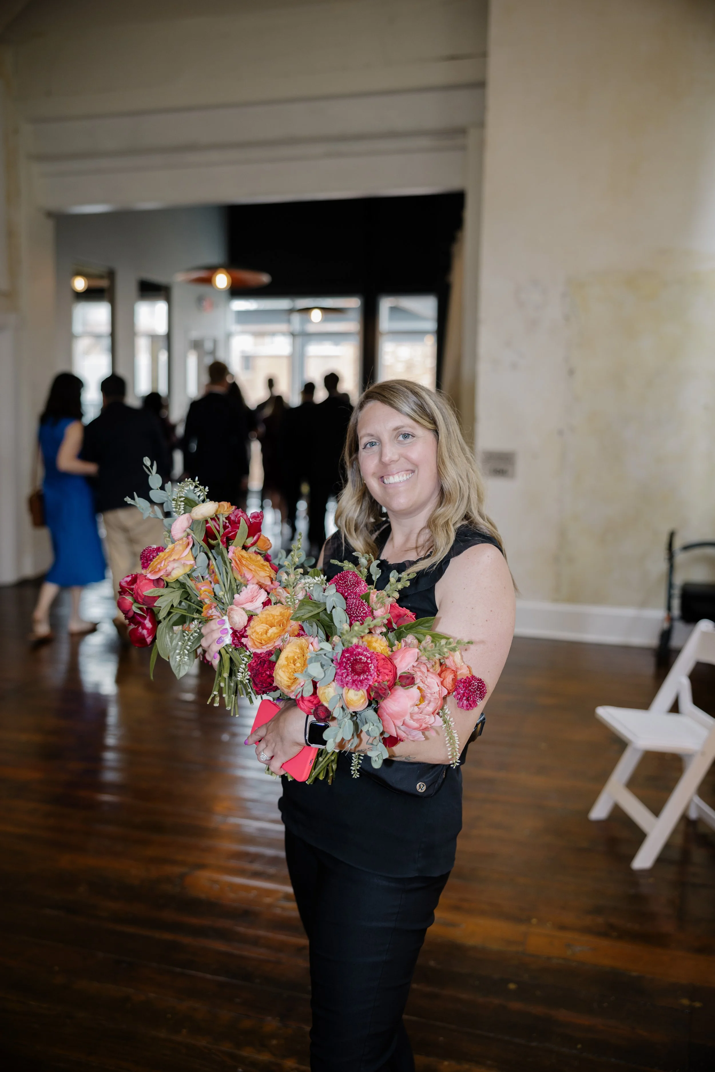 A woman smiling and holding a colorful bouquet of flowers indoors at a social event.