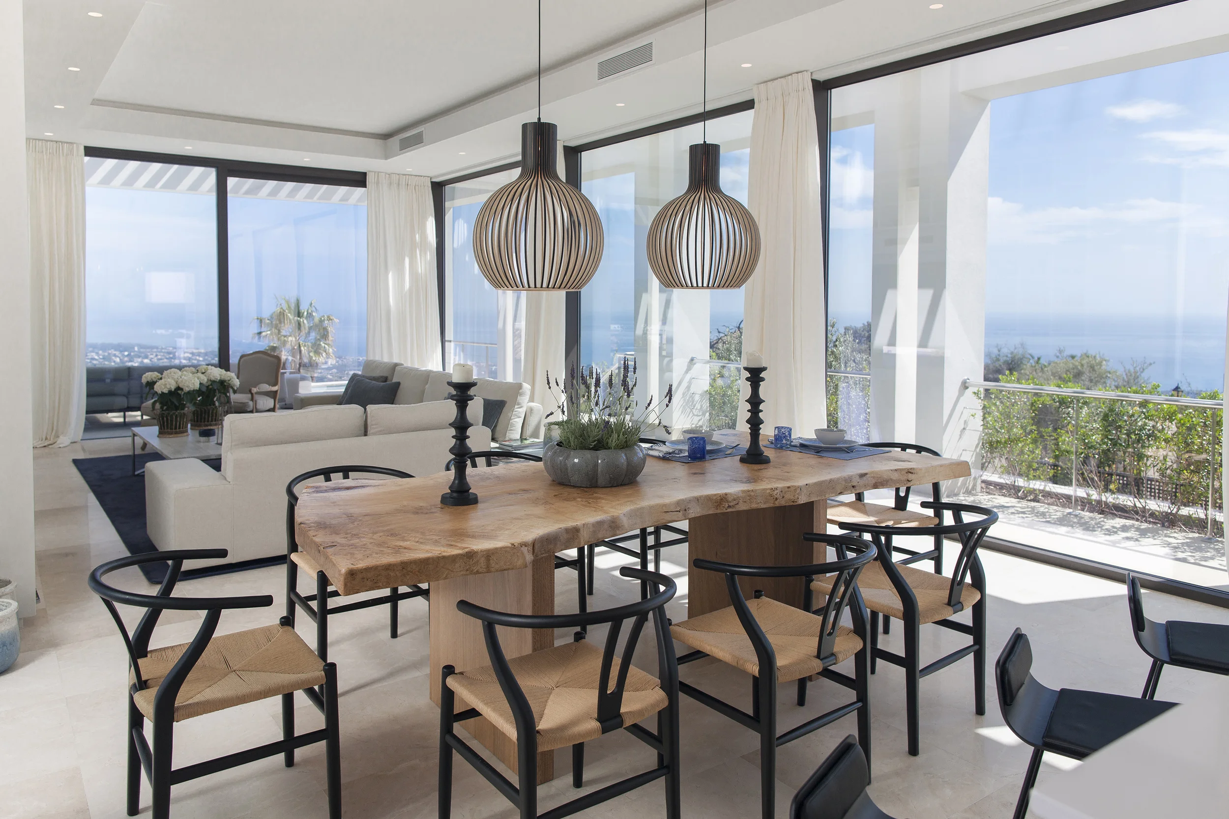Modern open-plan living and dining room with wooden table, black chairs, pendant lights, large windows, and ocean view.