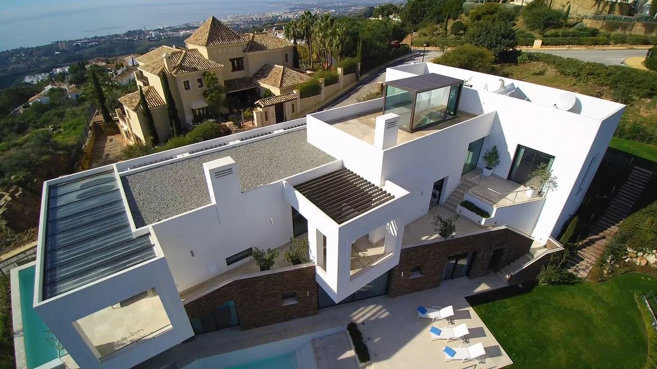Modern white villa with a pool, glass windows, and outdoor seating, set in a lush, landscaped area with views of other houses and the sea in the background.