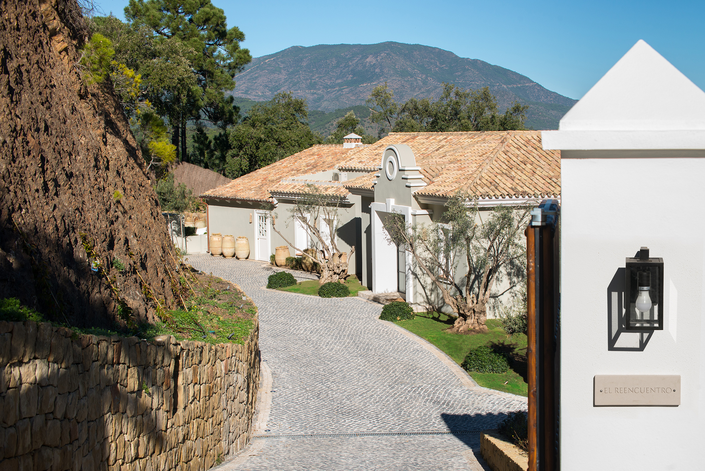 Mediterranean-style house with terracotta roof, cobblestone driveway, and surrounding natural landscape with mountains and trees.