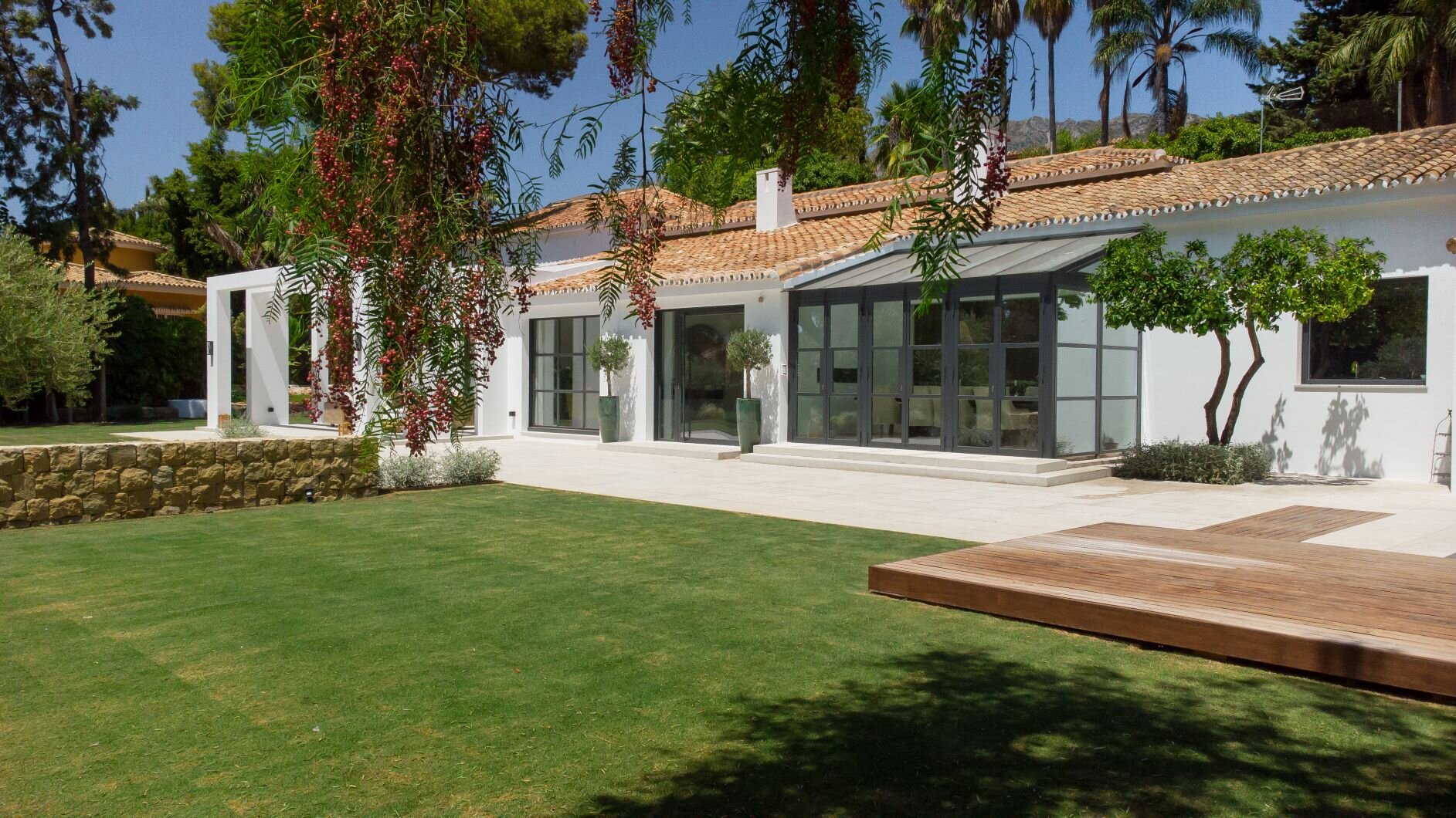 Modern white house with large windows, surrounded by a well-maintained lawn, trees, and plants. The house features a wooden deck and a tiled roof.