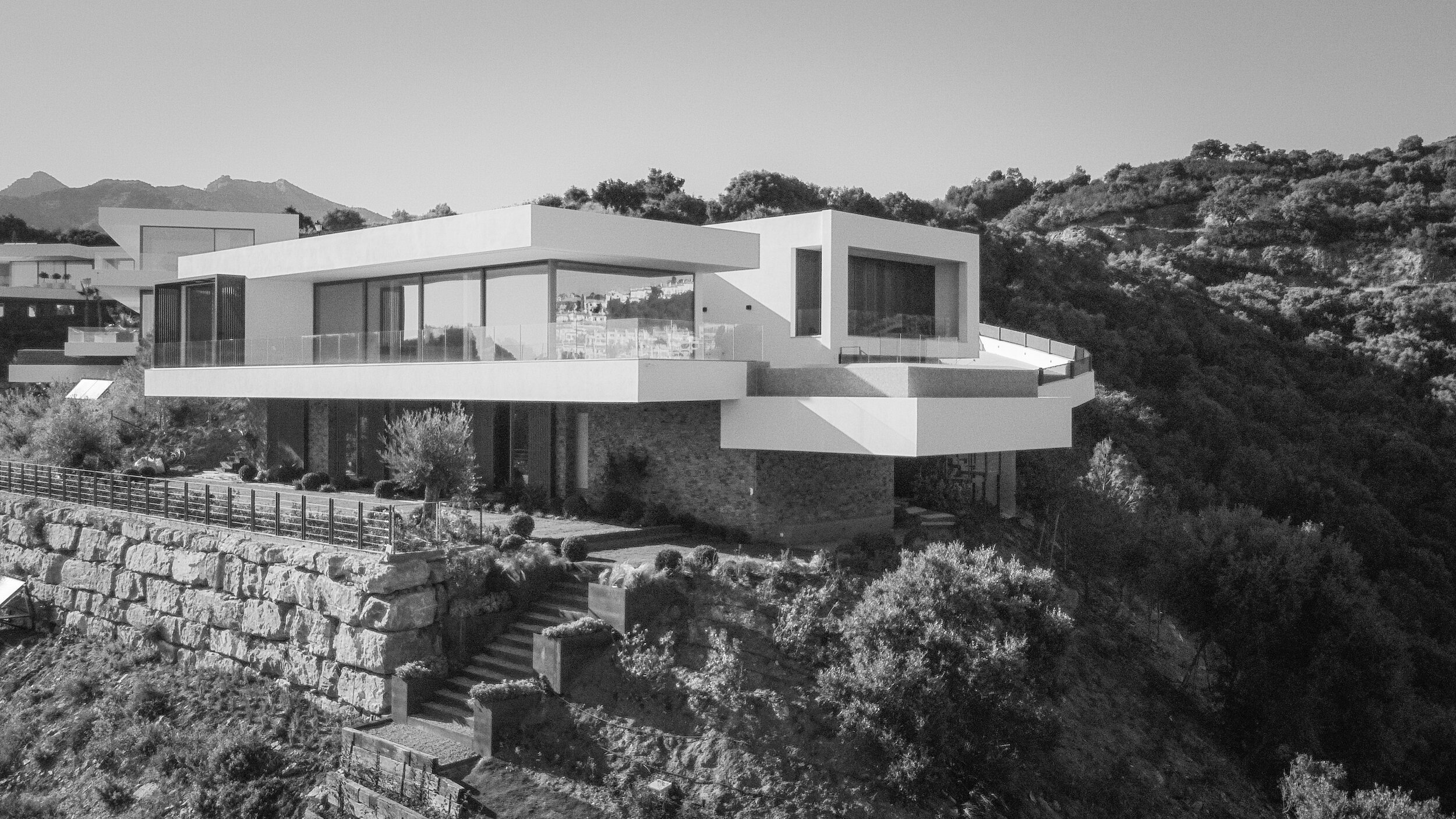 Modern hillside house with large windows and minimalist design, surrounded by trees and a stone retaining wall, set against a mountainous background.