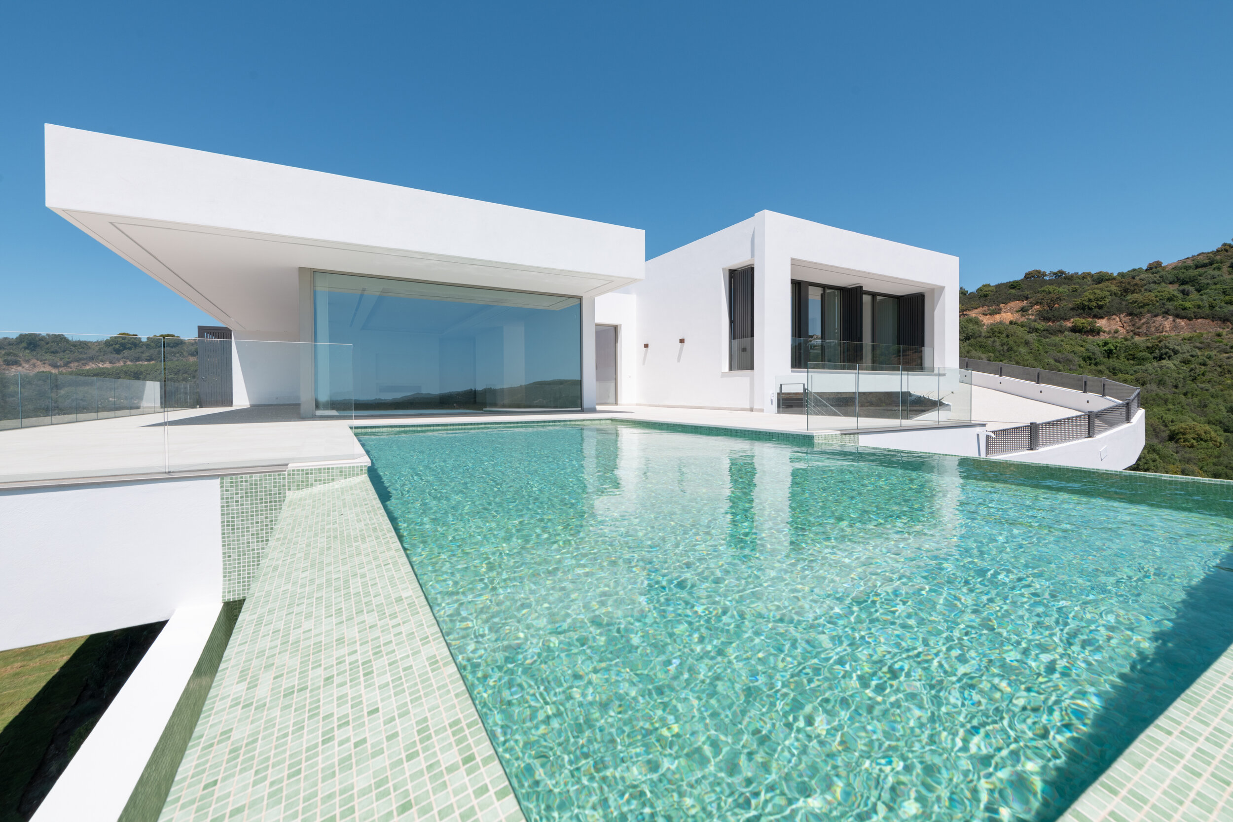 Modern white house with large glass windows and infinity pool overlooking green hills and clear blue sky.