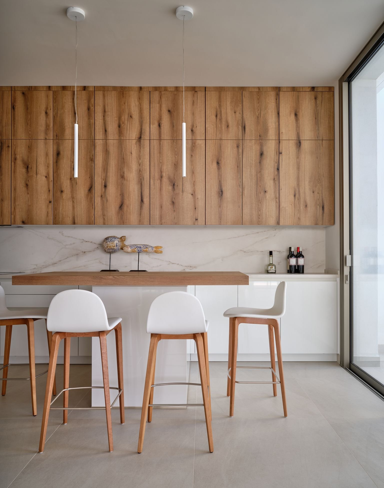 Modern kitchen with wooden cabinets, a white island with a wooden countertop, and white bar stools. Pendant lights hang from the ceiling, and there is a decorative fish figure on the counter. Bottles are placed in the corner.