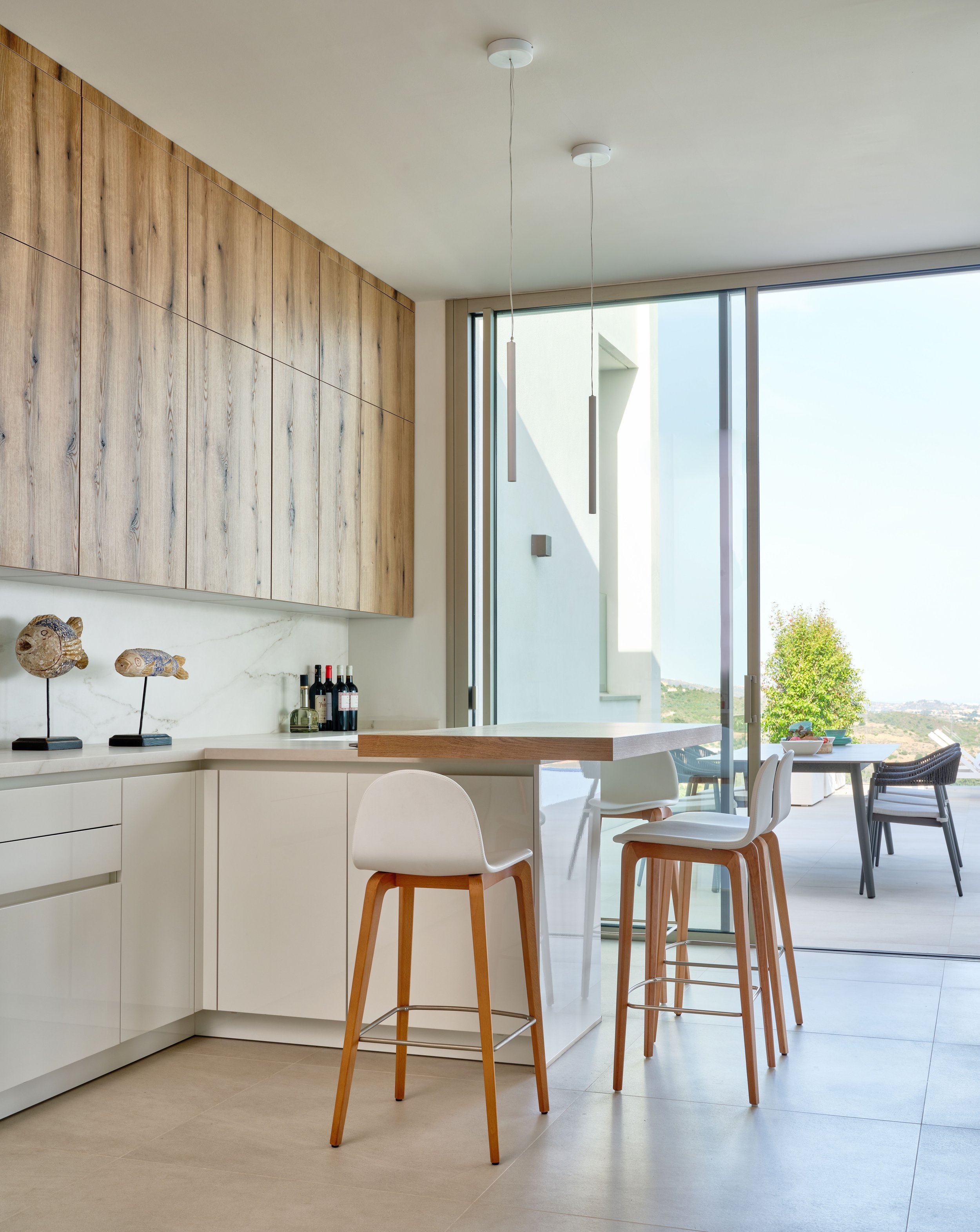 Modern kitchen with white cabinets, wooden accents, bar stools, pendant lights, and a large glass door leading to a patio with outdoor seating. Sculptures and wine bottles are on the counter.
