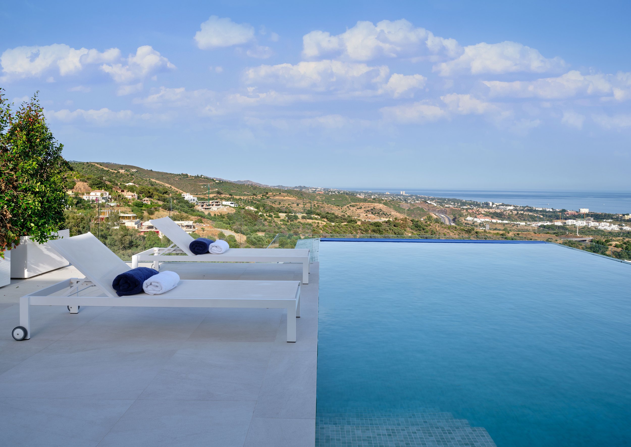 Infinity pool with lounge chairs and towels overlooking hilly landscape and distant city.