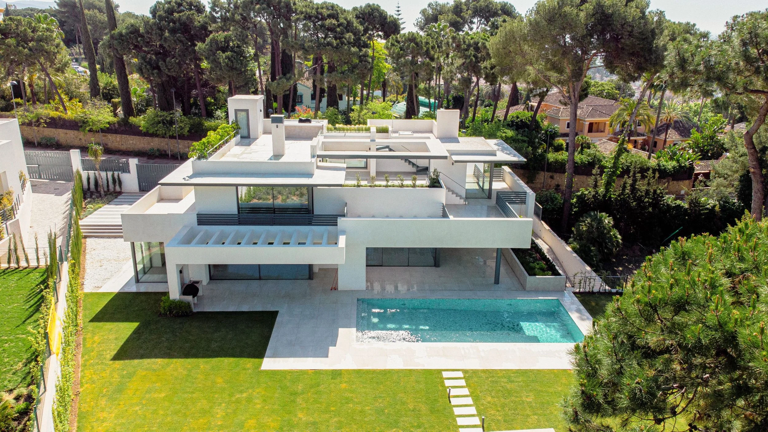 Modern white villa with a flat roof, landscaped garden, and swimming pool, surrounded by trees.