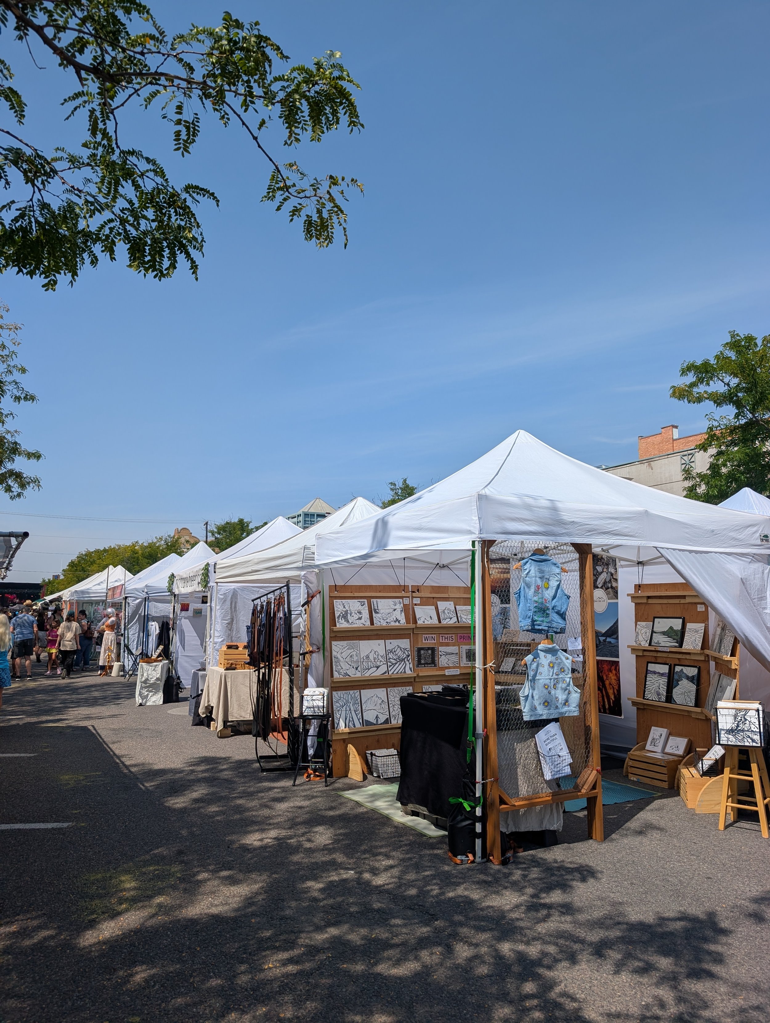 a row of art market booths on a street with shadows from a nearby tree