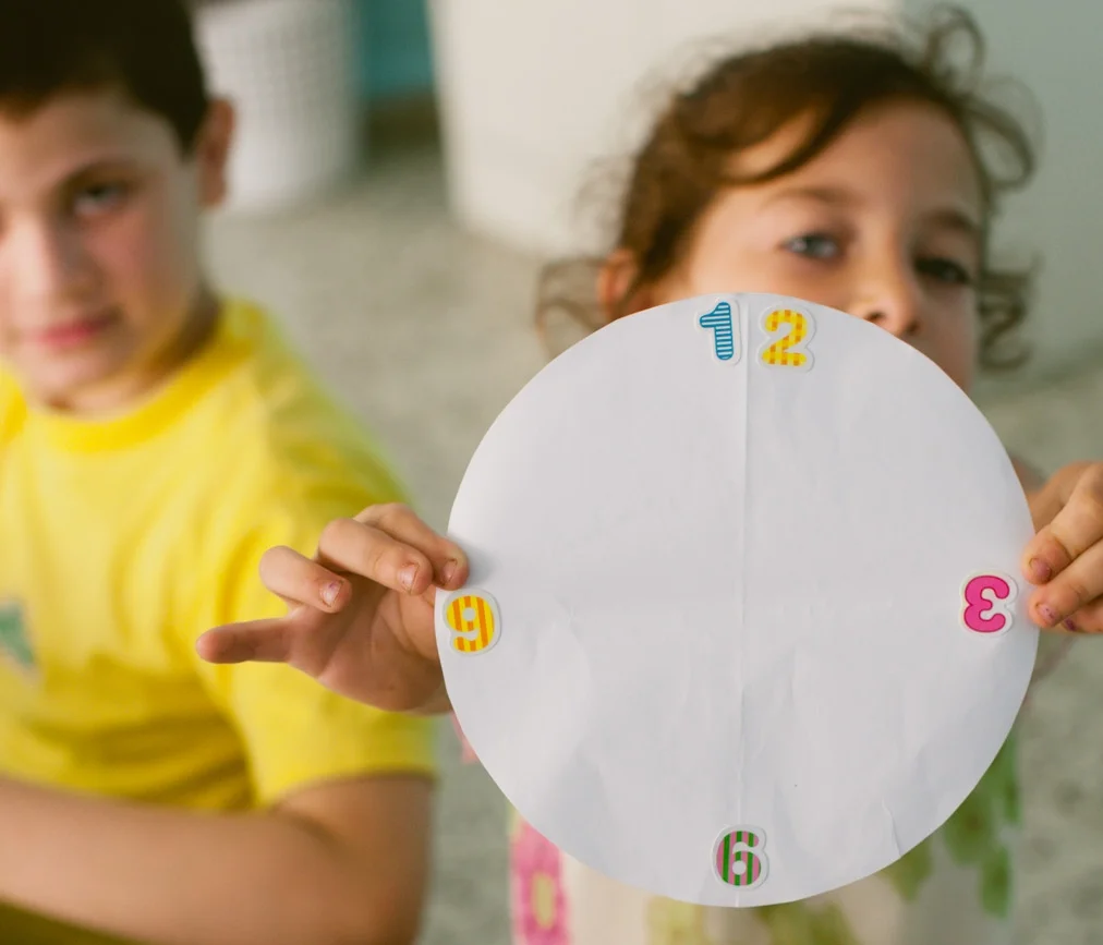 A girl displays her art project, which resembles a clock, while a boy in the left background looks on.