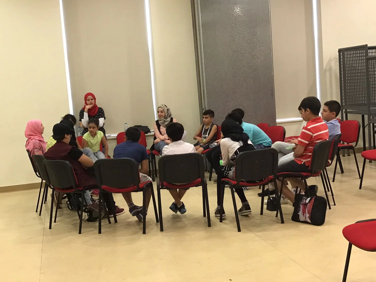 In the picture: a group of about fourteen students sit on chairs around a table, with two teachers (one standing, one sitting in a chair), and discuss "Symbolism and Storytelling."