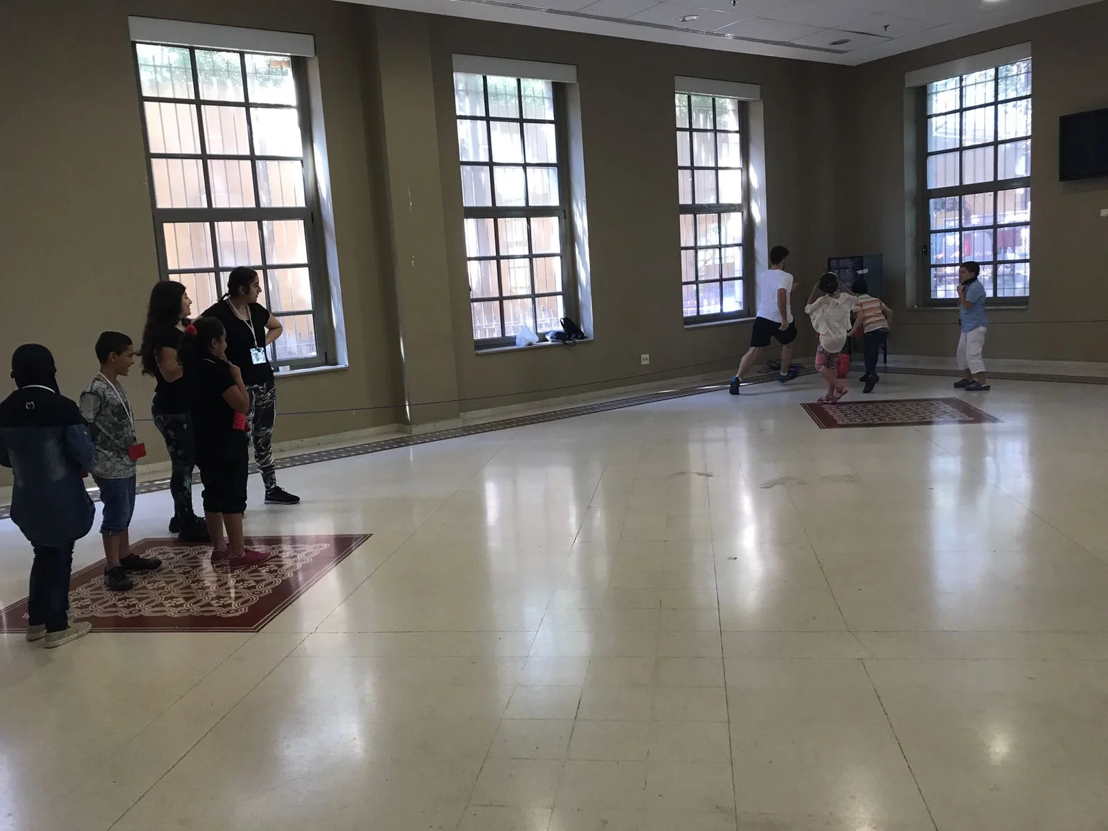 In the picture: a group of about four children, to the left, watch a group of about four other children, to the right, practice a soccer drill in the corner of an open room while a volunteer supervises.