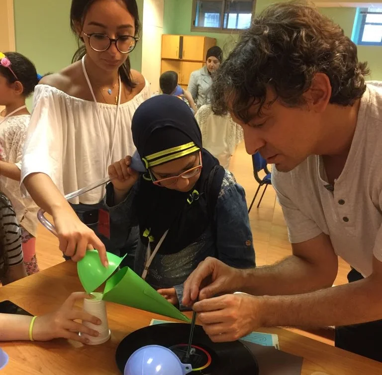 In the picture: Tim Mauro, far right, assists a student (center) and volunteer (far left) with putting together a record player made of paper, a pin, a pencil, and tape during a science class.