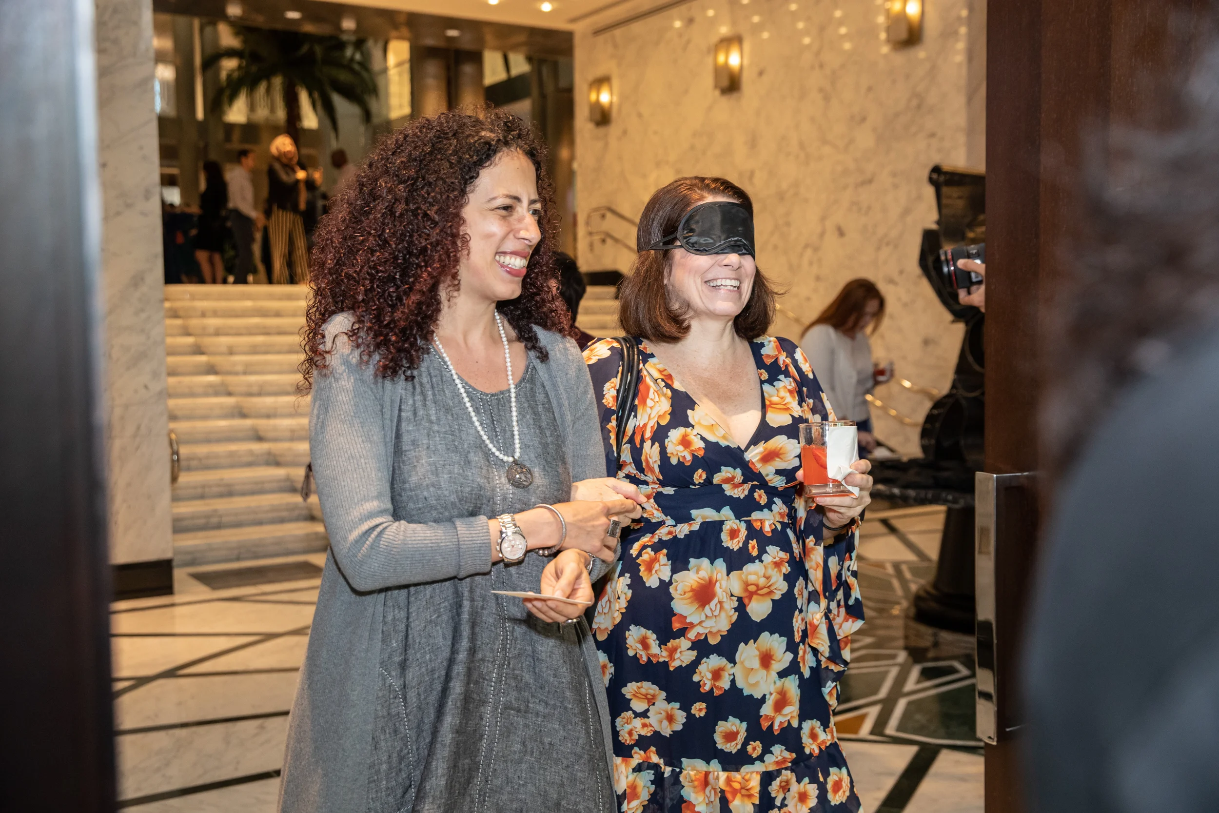 A sighted guide leads a blindfolded participant to their seat. Both women are laughing.