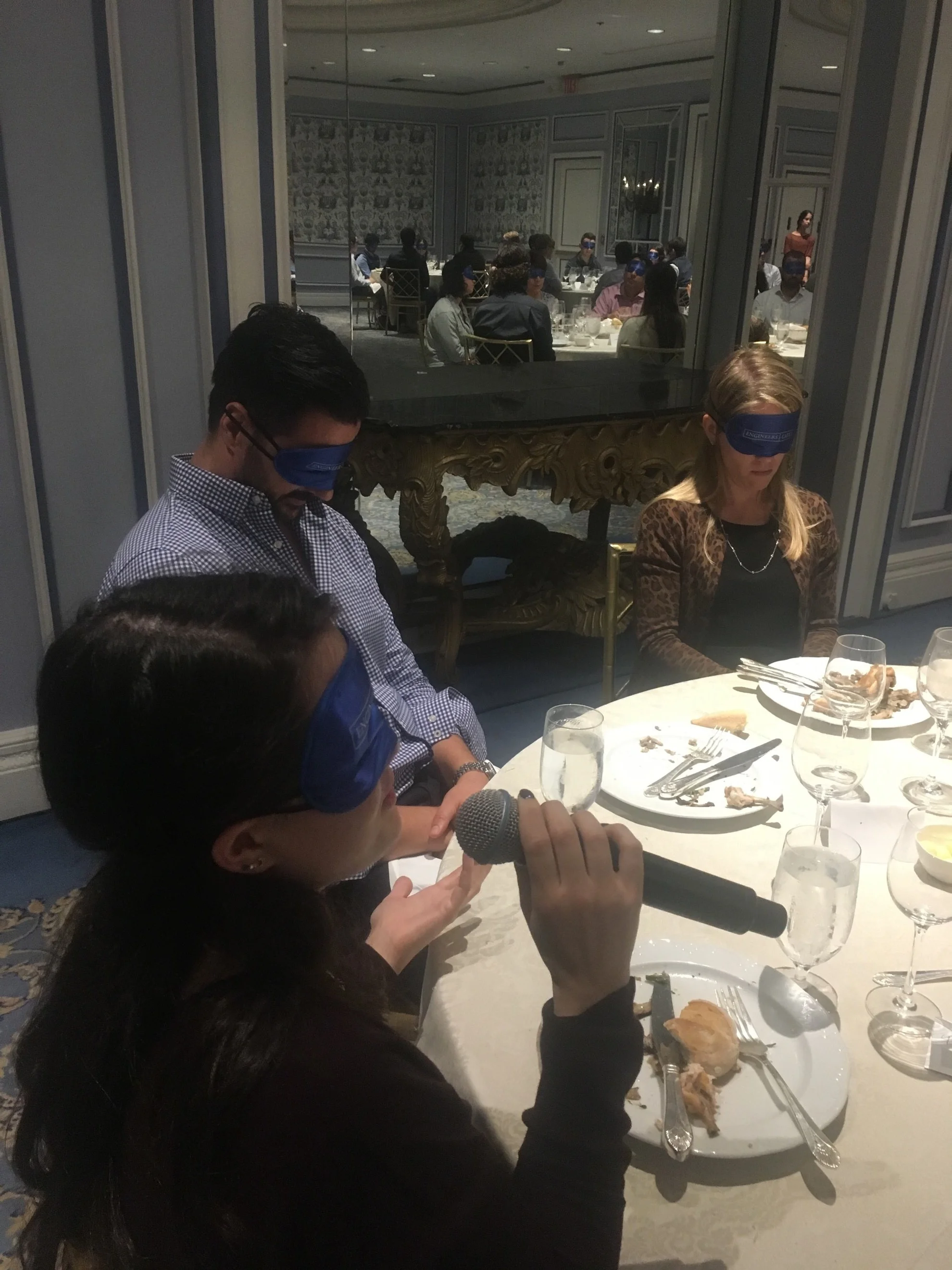 Blindfolded woman sitting at a table answering a question while other blindfolded participants are listening