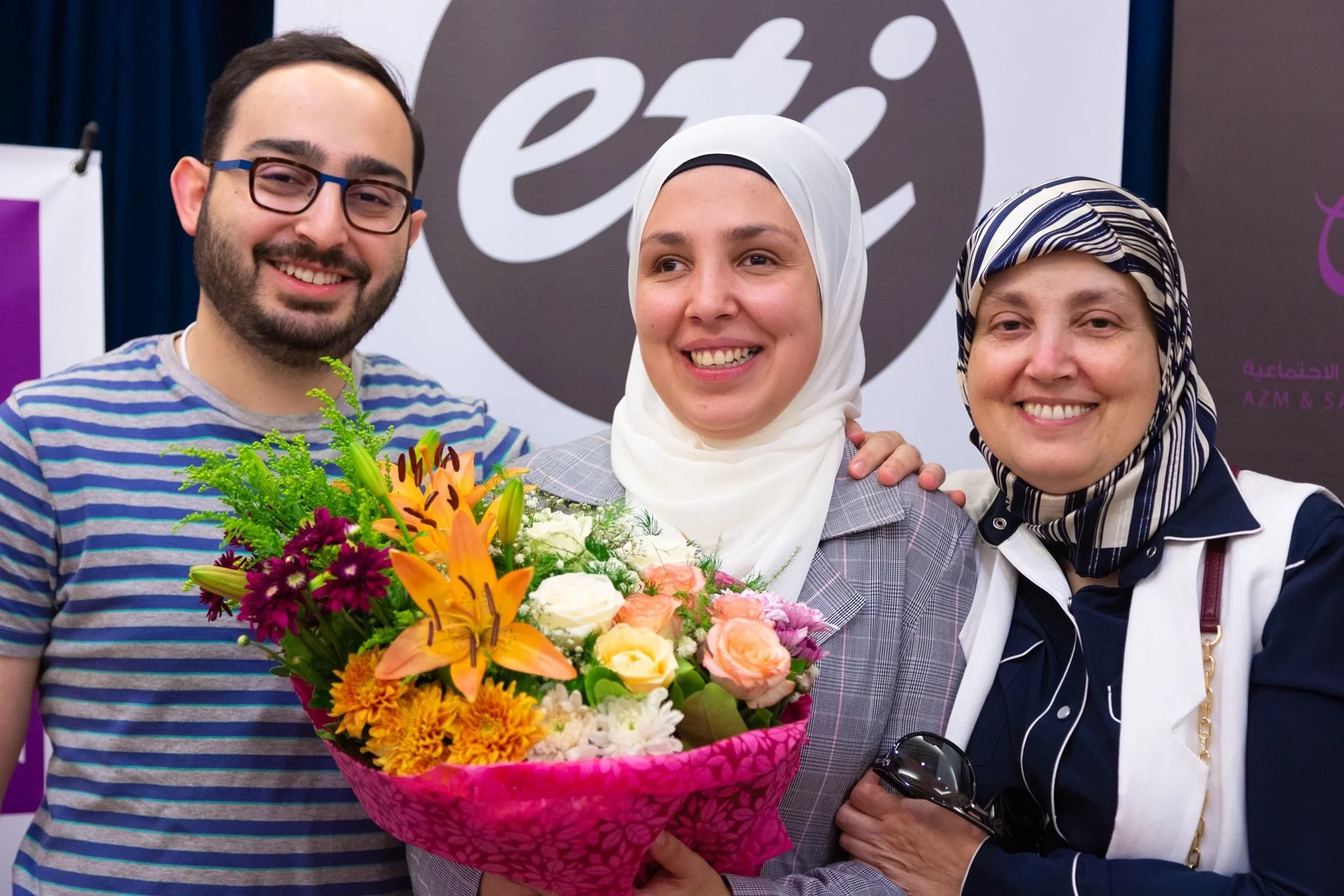 Two women and a man stand in front of a sign that reads “ETI” All three are smiling brightly. The woman in the middle holds a bright bouquet of flowers, The vibrant flowers illustrate the affection and pride that Sara’s family has for her.