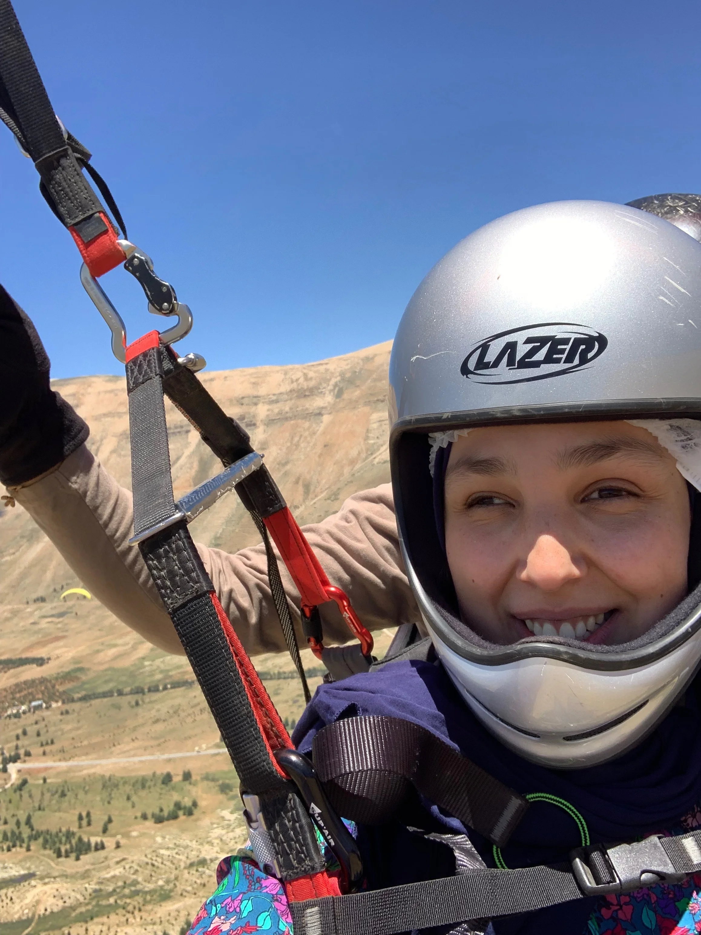 A close up image shows a woman smiling at the camera, wearing a silver helmet and with straps and clips covering her chest. Behind her we see the hand of a guide controlling levers. The background is a beautiful valley.