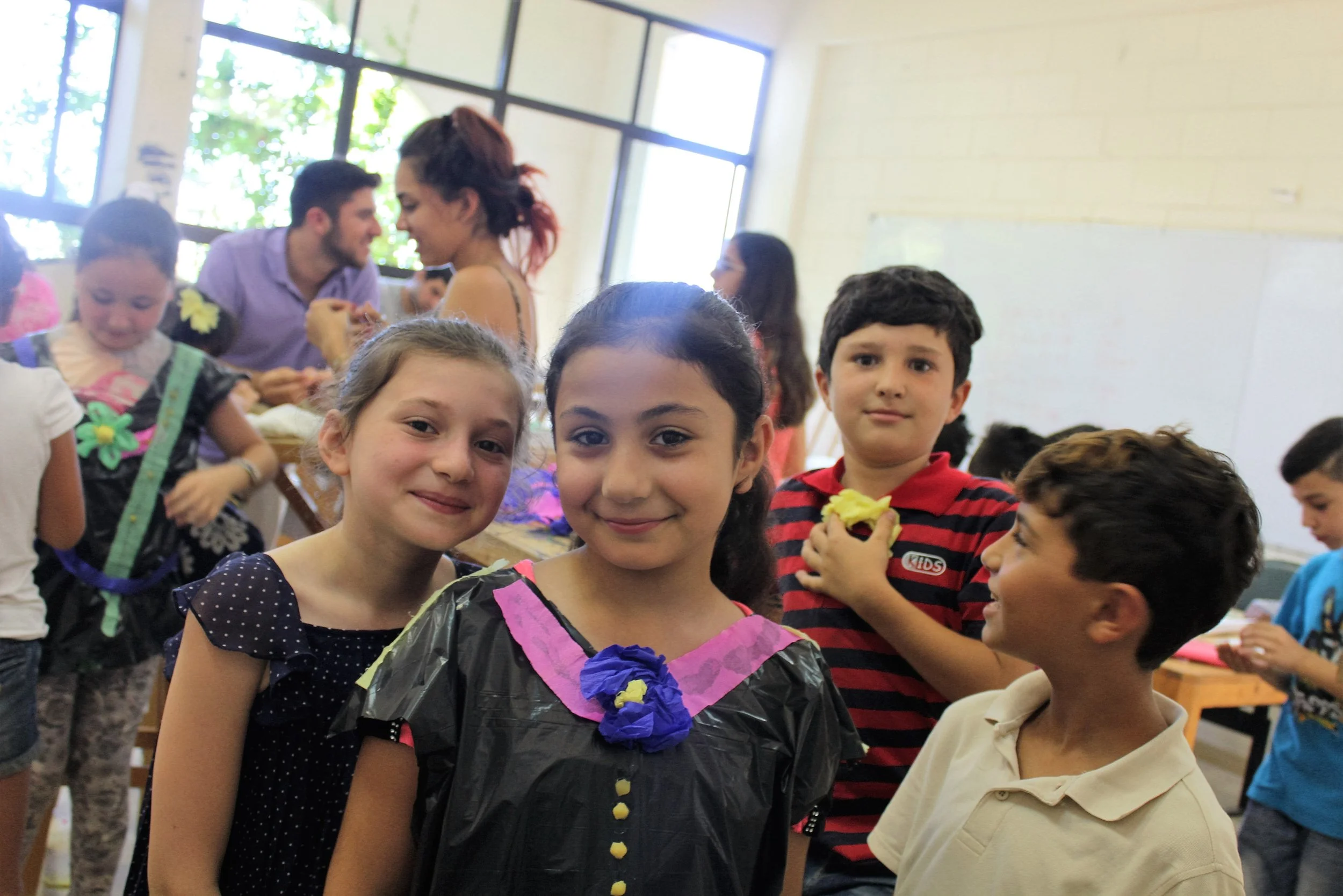 Two young girls face the camera, smiling. In the background, other children are talking and playing.