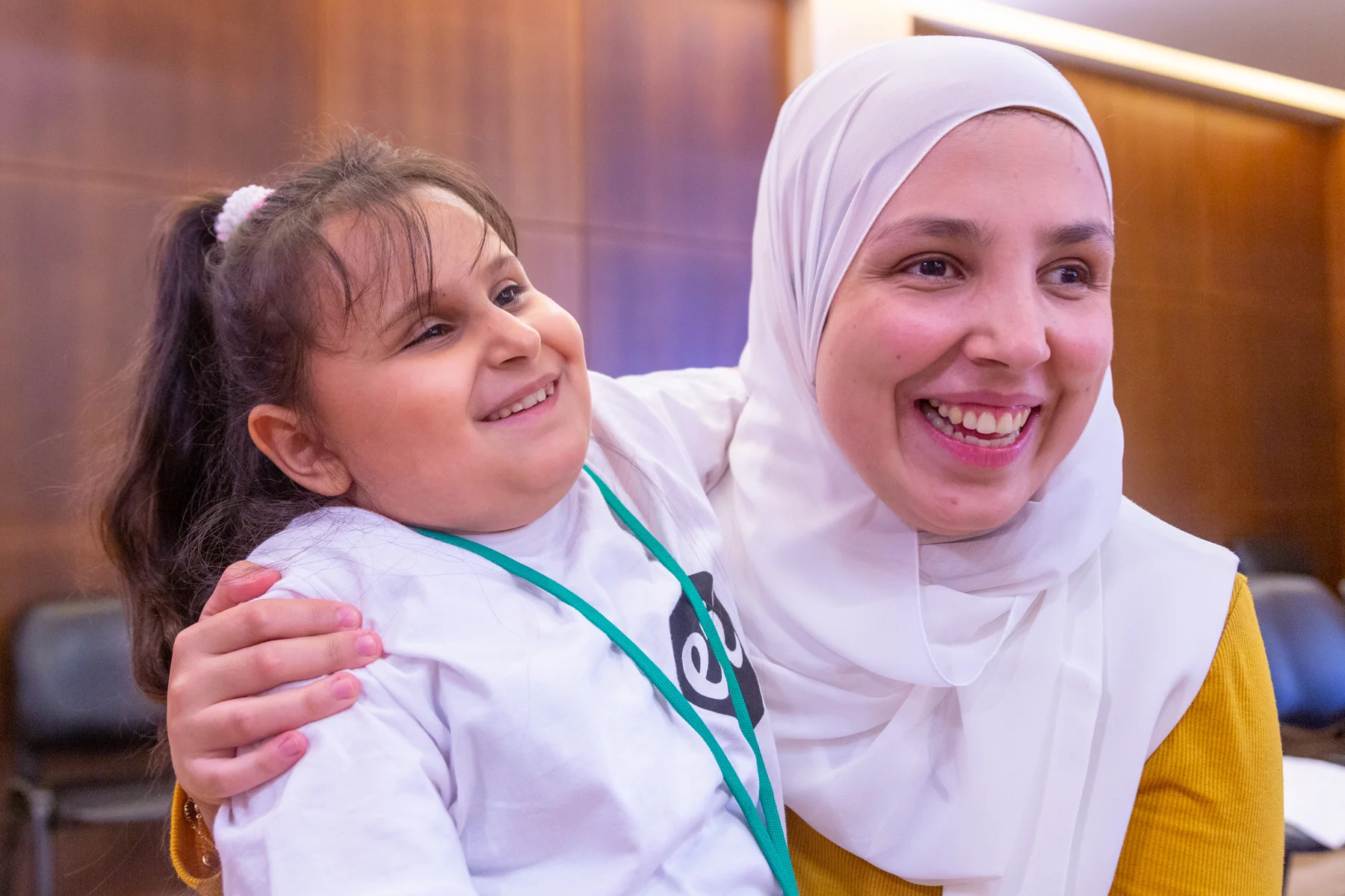 A young girl with a brown ponytail wears a white t-shirt with the ETI logo in the upper left-hand corner. Only the letter “e” is completely visible as the girl is wearing a turquoise lanyard. She has her left arm stretch around the next of a woman in a white hajib and bright yellow shirt. Both woman and child are smiling