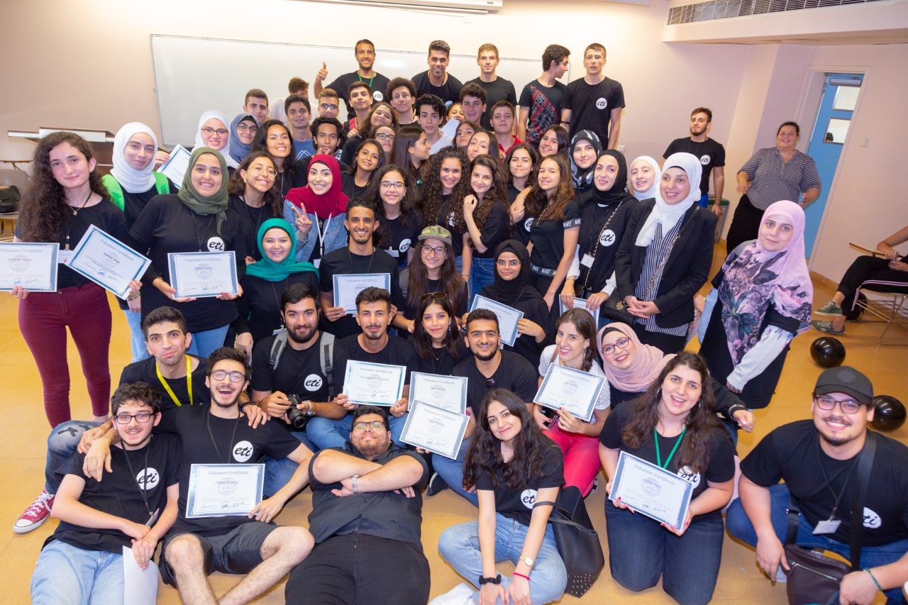 A large group of people in black t-shirts are grouped in front of the camera, smiling and holding white pieces of paper with blue borders.