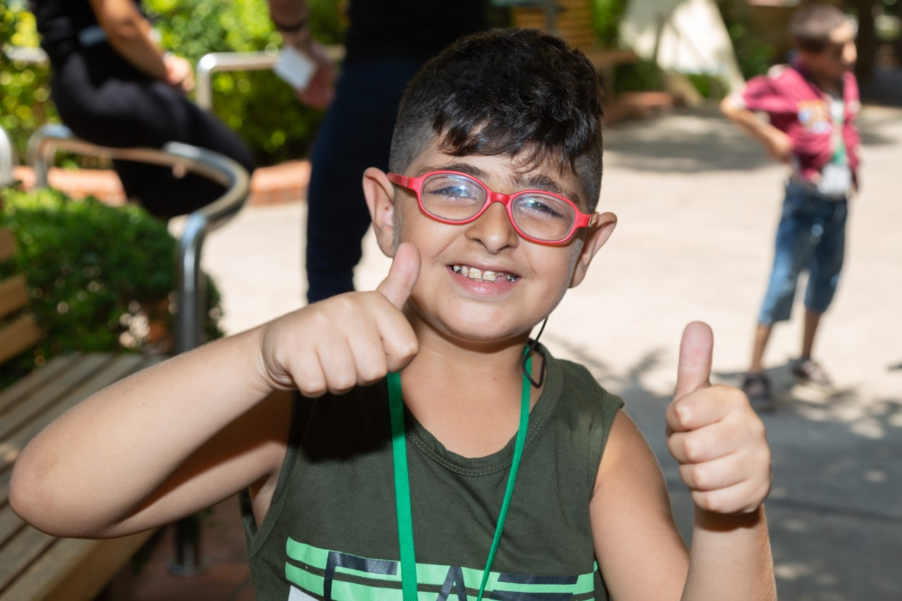 A boy with red glasses holds two thumbs up. He has black hair and is wearing a green tank top.