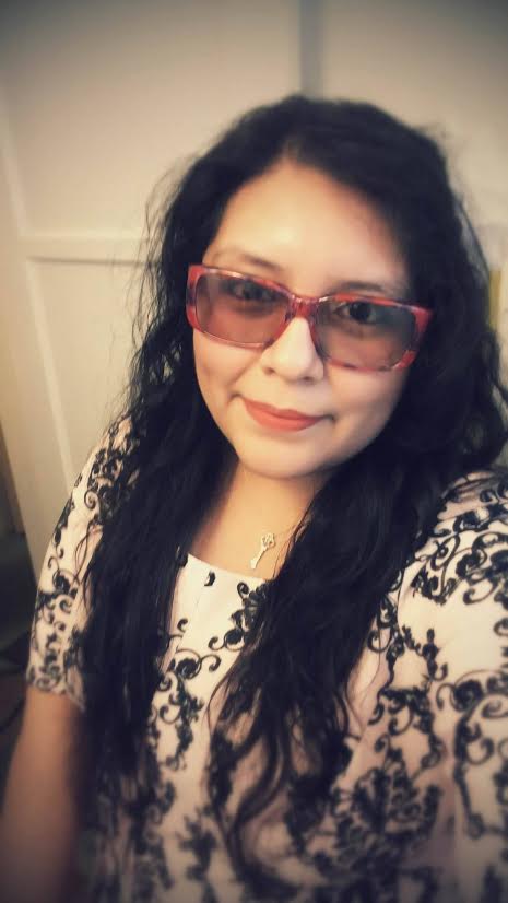 a photo of a young woman with long dark hair wearing red framed glasses and smiling slightly. The corners of the photo are blurry.