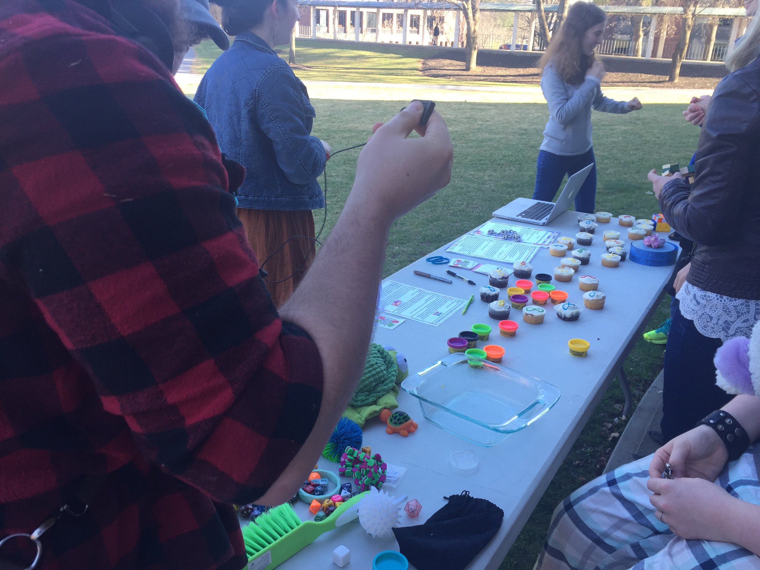 Image description: Cropped view of people standing at a table displaying autism acceptance materials, mini tins of Play-do, cupcakes and stim toys.