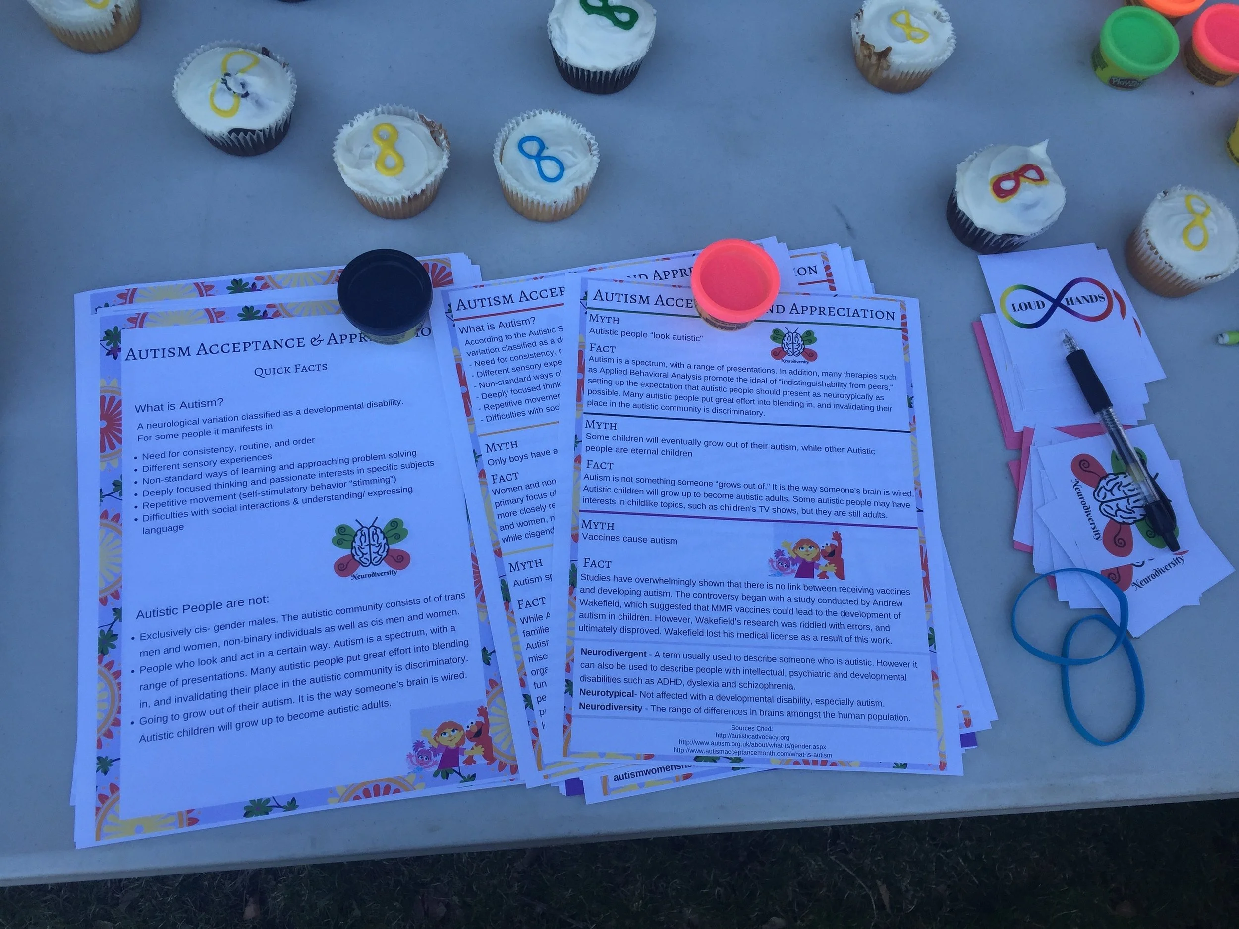 Image description:: Close up view of autism acceptance flyers, cupcakes with colorful infinity symbols on white frosting and mini tins of Play-do