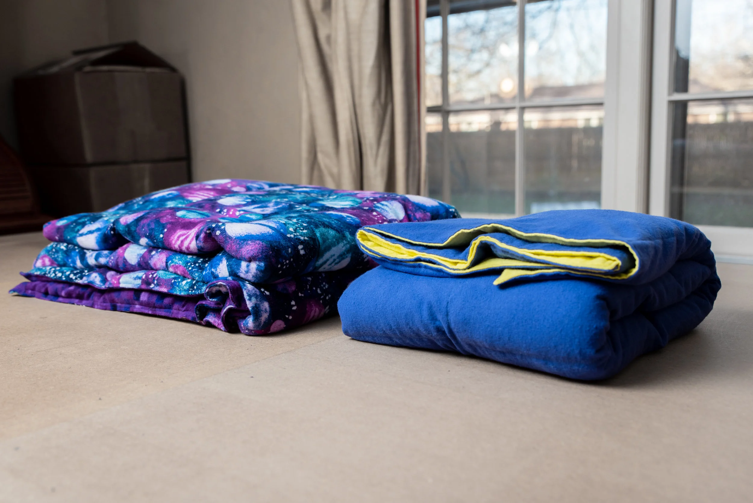 Two weighted blankets folded side by side on a bed. The one on the left has a fabric pattern of deep blues, purples and white and the one on the right is two toned, with blue on the outer side and yellow on the side that is folded inward.