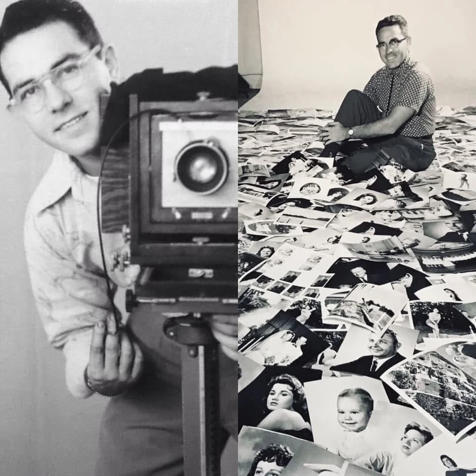 A black and white photo of a man sitting cross-legged on the floor, surrounded by numerous photographs of people. The man is smiling and wearing glasses, a checkered shirt, and dark pants.