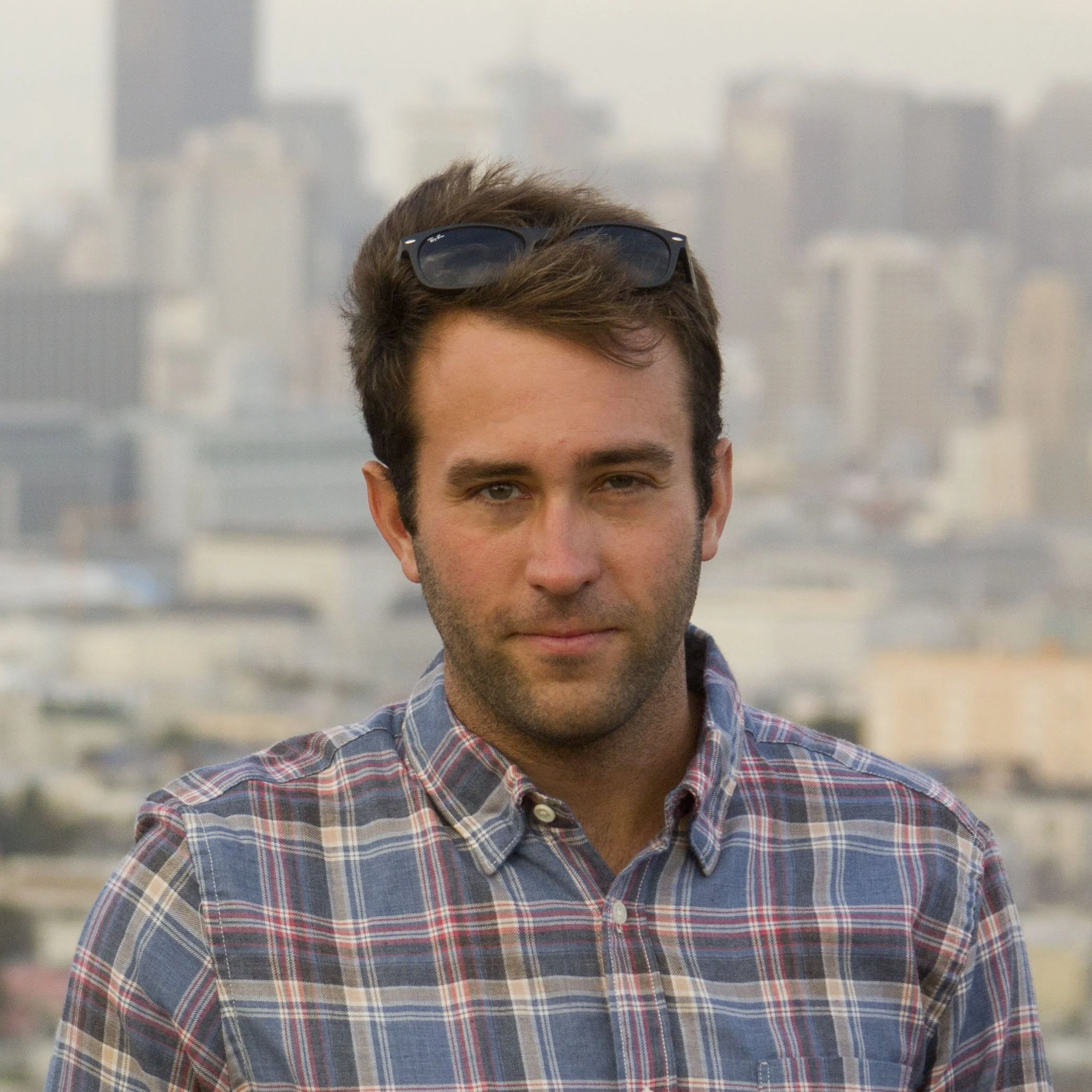 Close-up of a young man with brown hair, wearing sunglasses on his head and a plaid shirt, standing outdoors in a city.