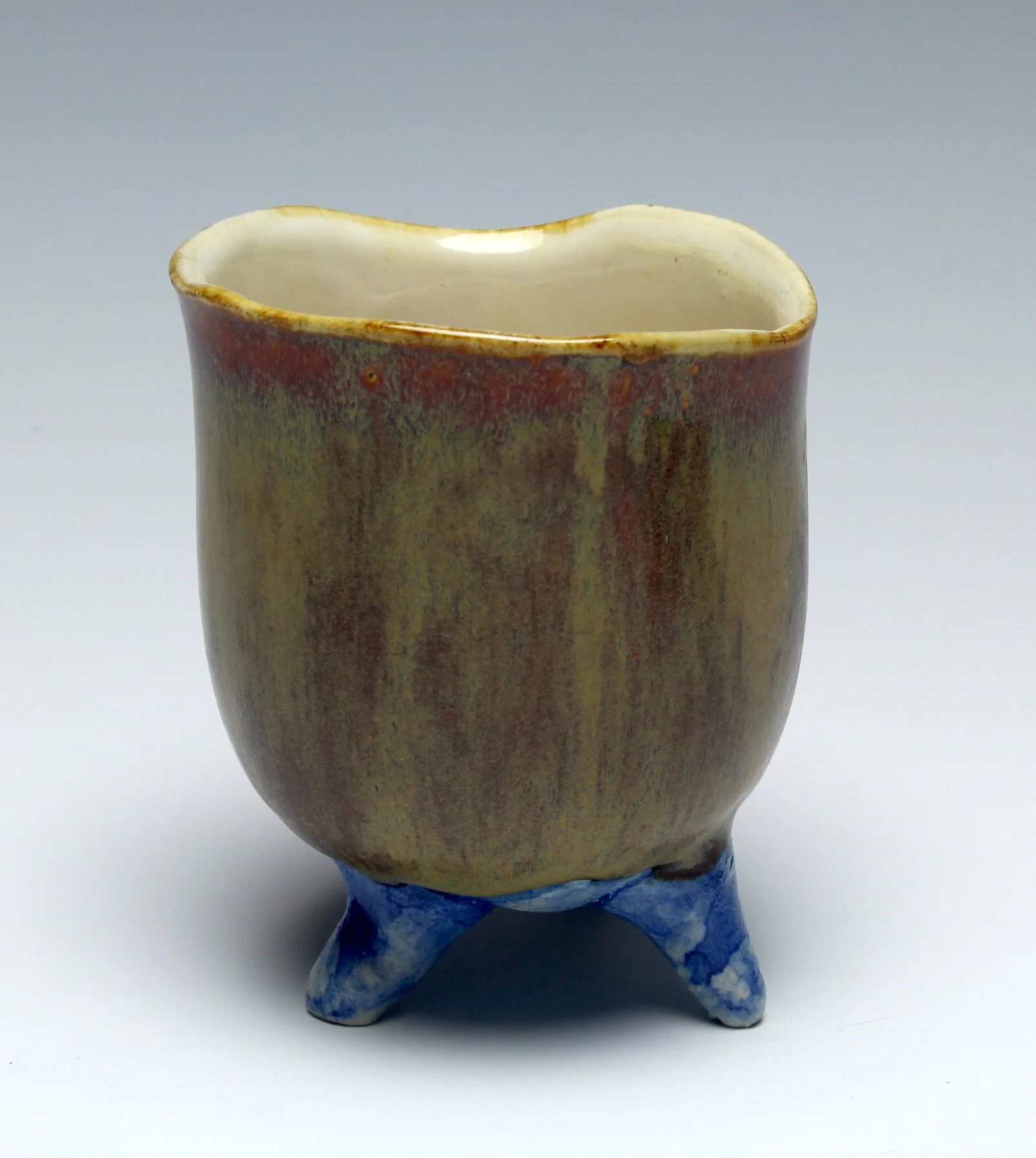 A small cup with 4 little blue feet, it is shaped like a tulip. The cup is reddish tan with an ivory interior.