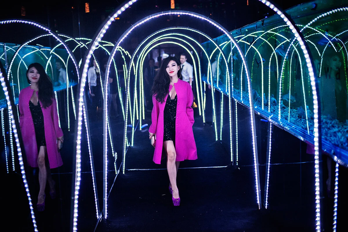 October 25, 2013, Shanghai, China -  A female guest dressed in a sparkly short black dress, mauve coat, mauve heels and bright red lipstick  strides down a corridor lined with a long tank filled with small sharks and decorated with white neon arches