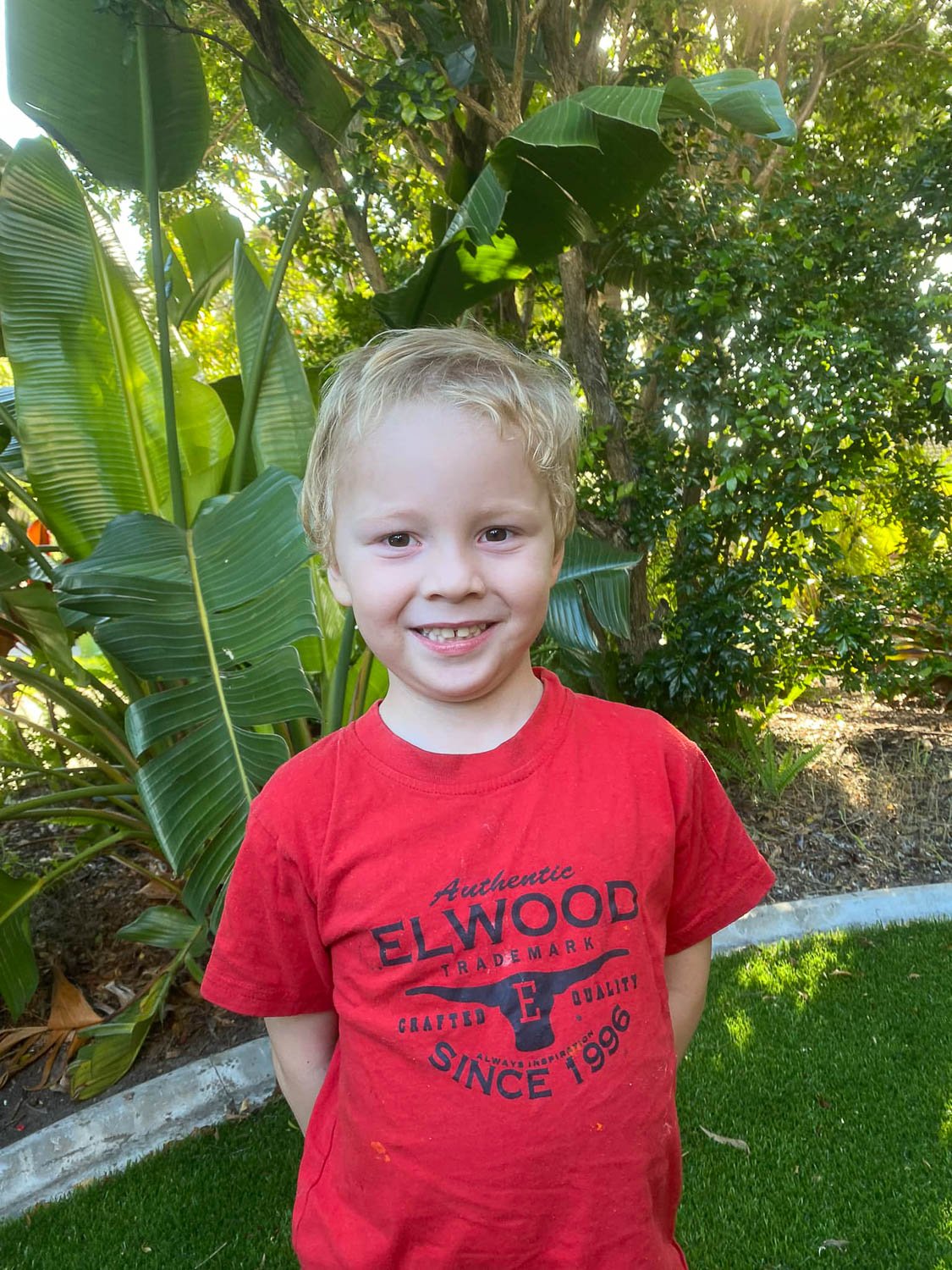 A blonde haired young boy in a red t-shirt standing in front of leafy background