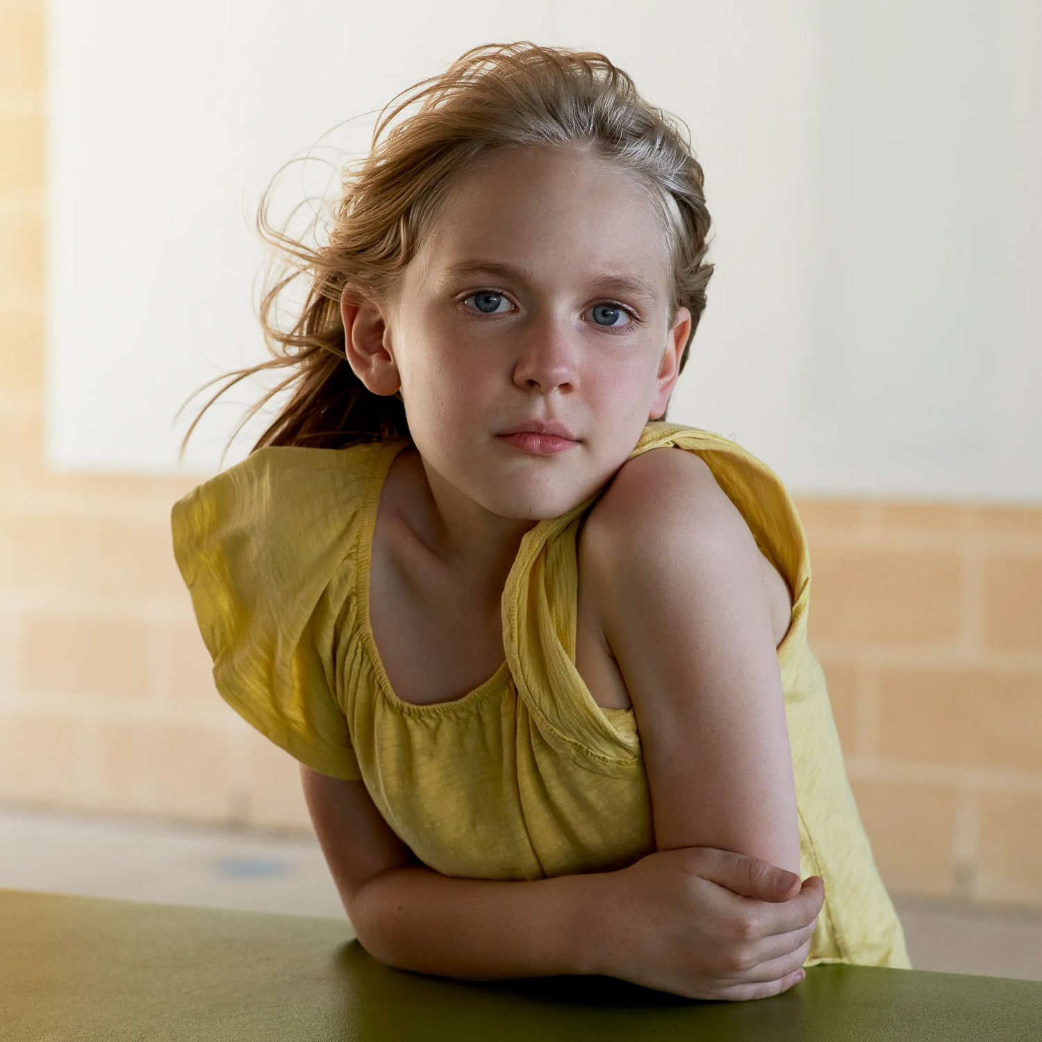 A young blonde girl in a yellow dress leaning on a table looking at camera
